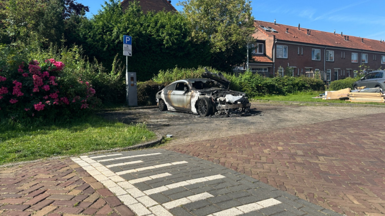 Verbrande auto geparkeerd op een stoep in een woonwijk, omgeven door groene struiken en rozenstruiken, met rode bakstenen huizen op de achtergrond.