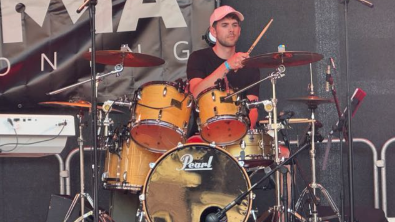 Een drummer met een roze pet speelt op een Pearl drumstel tijdens een live optreden op een podium.