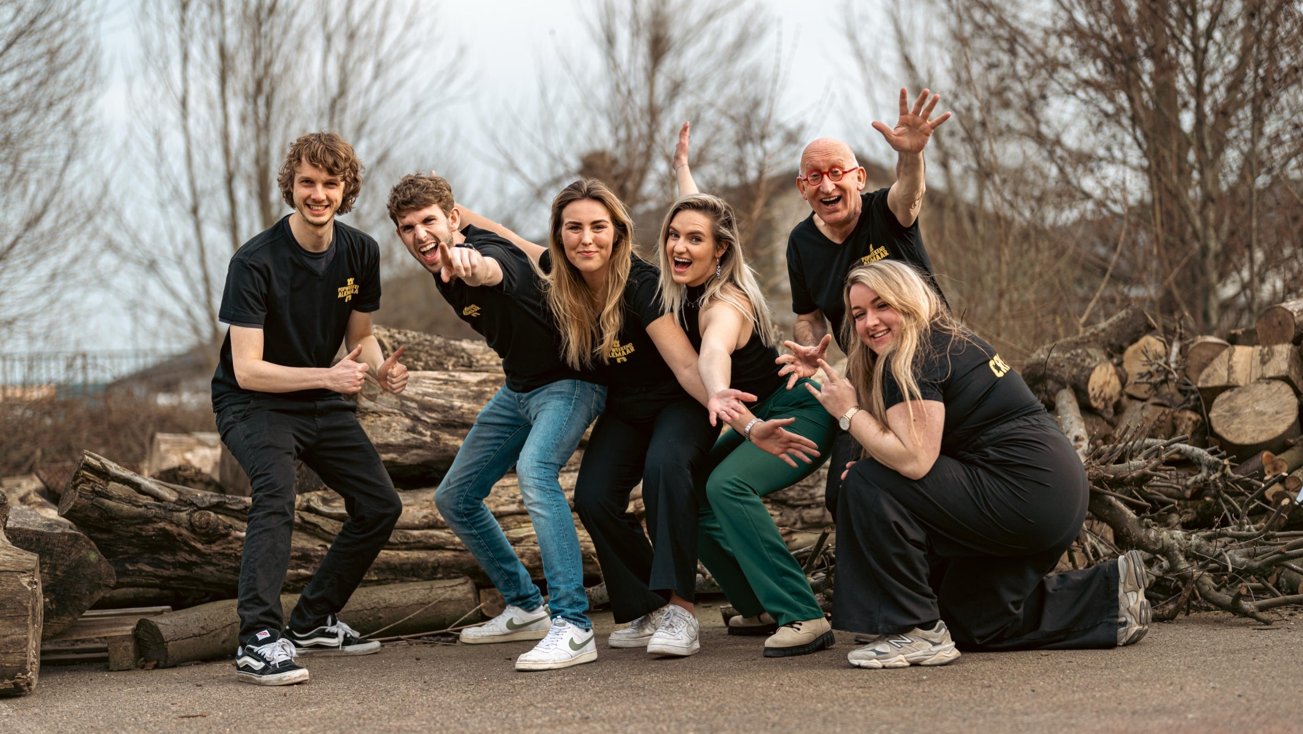 Een groep van vijf mensen poseert vrolijk buiten bij een stapel boomstammen, gekleed in zwarte shirts en casual kleding, met een grijze lucht en kale bomen op de achtergrond.