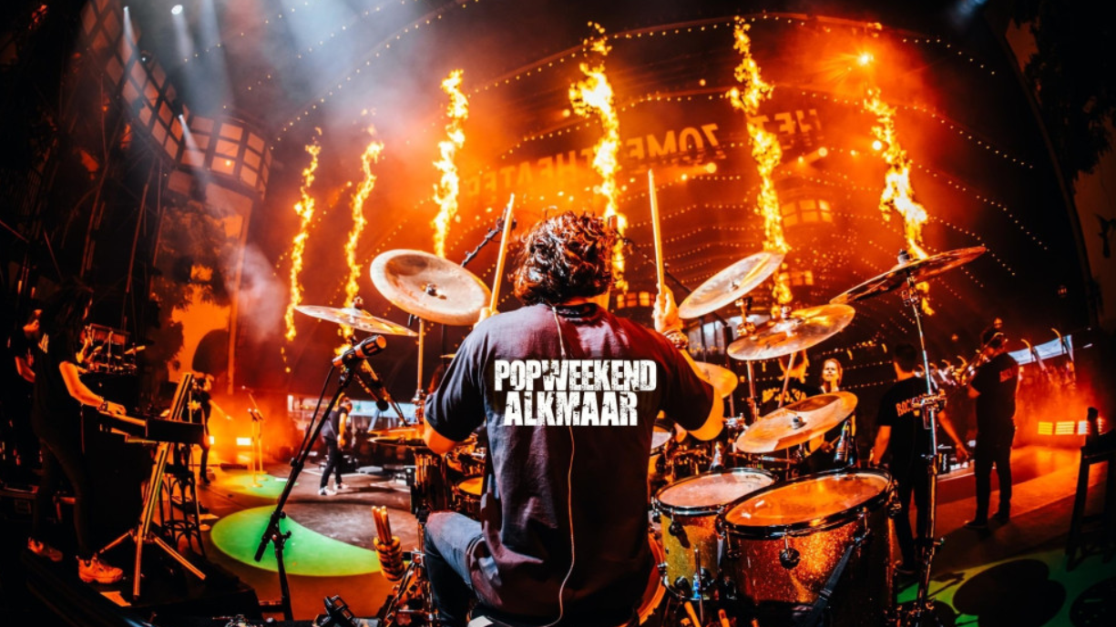 Een drummer speelt op een podium bij Popweekend Alkmaar, met vurige effecten en fel verlichte achtergrond.
