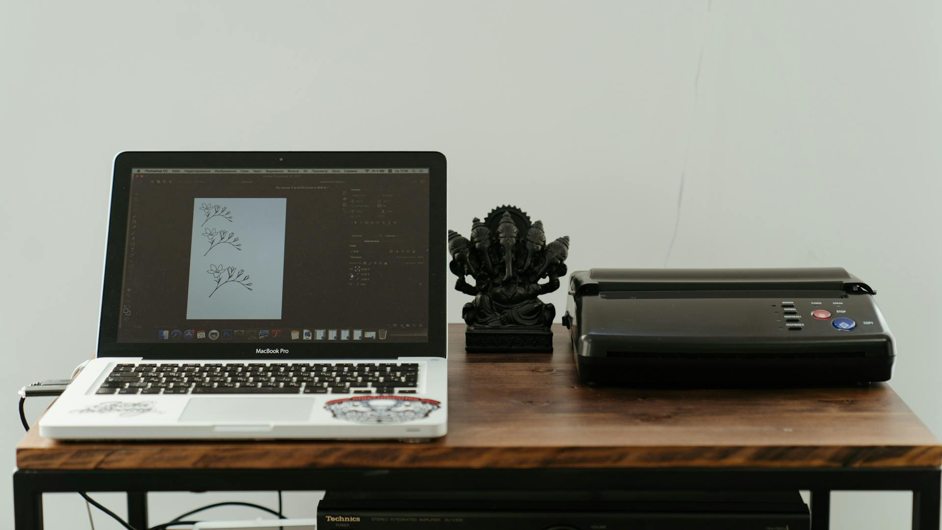 Een bureau met een opengeklapte MacBook Pro waarop een tekening van bloemen te zien is, naast een zwart beeldje en een printer.