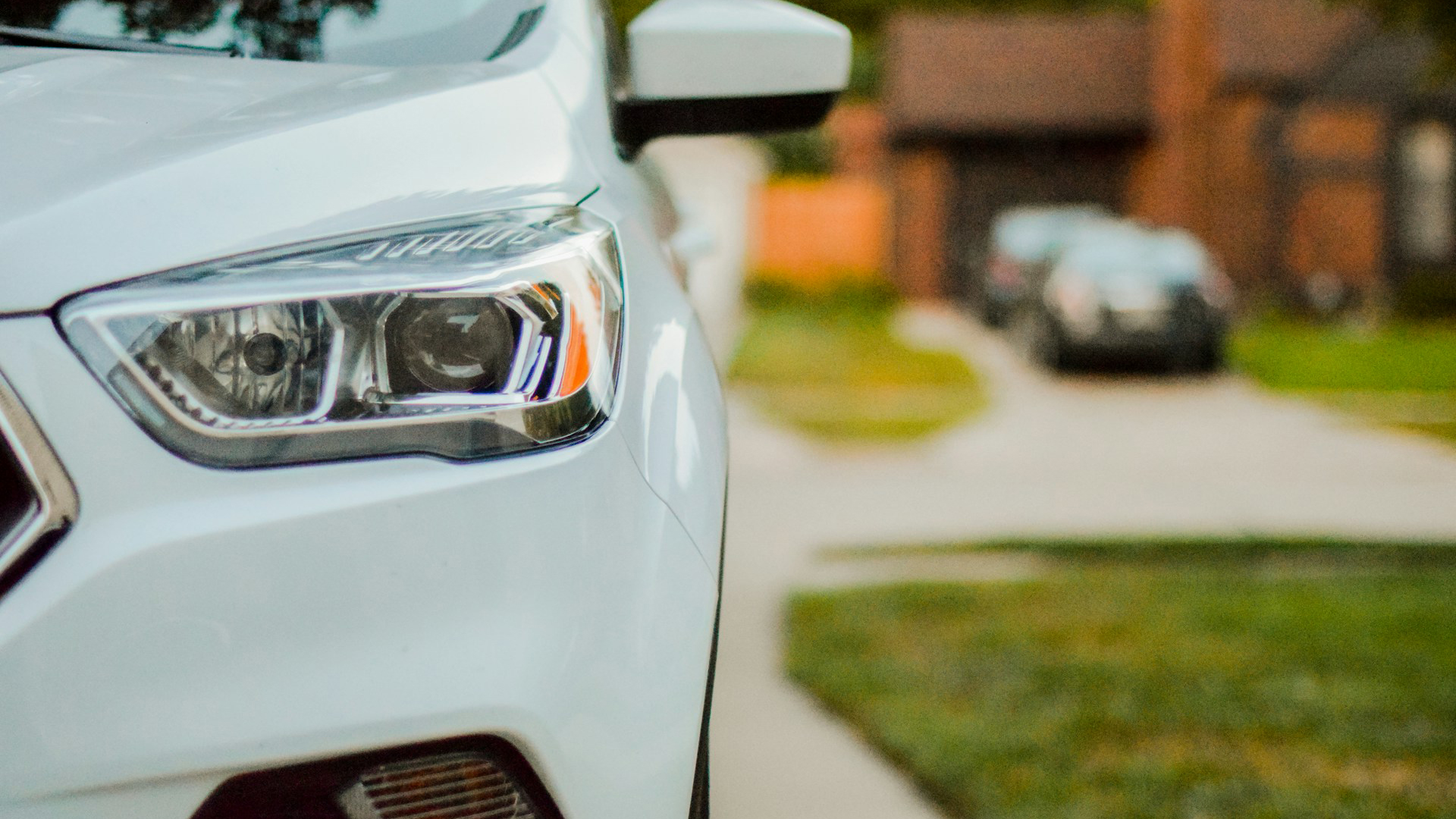 Vooraanzicht van een witte auto met focus op de rechterkoplamp, geparkeerd op een oprit met een ander voertuig en een bakstenen huis op de achtergrond.