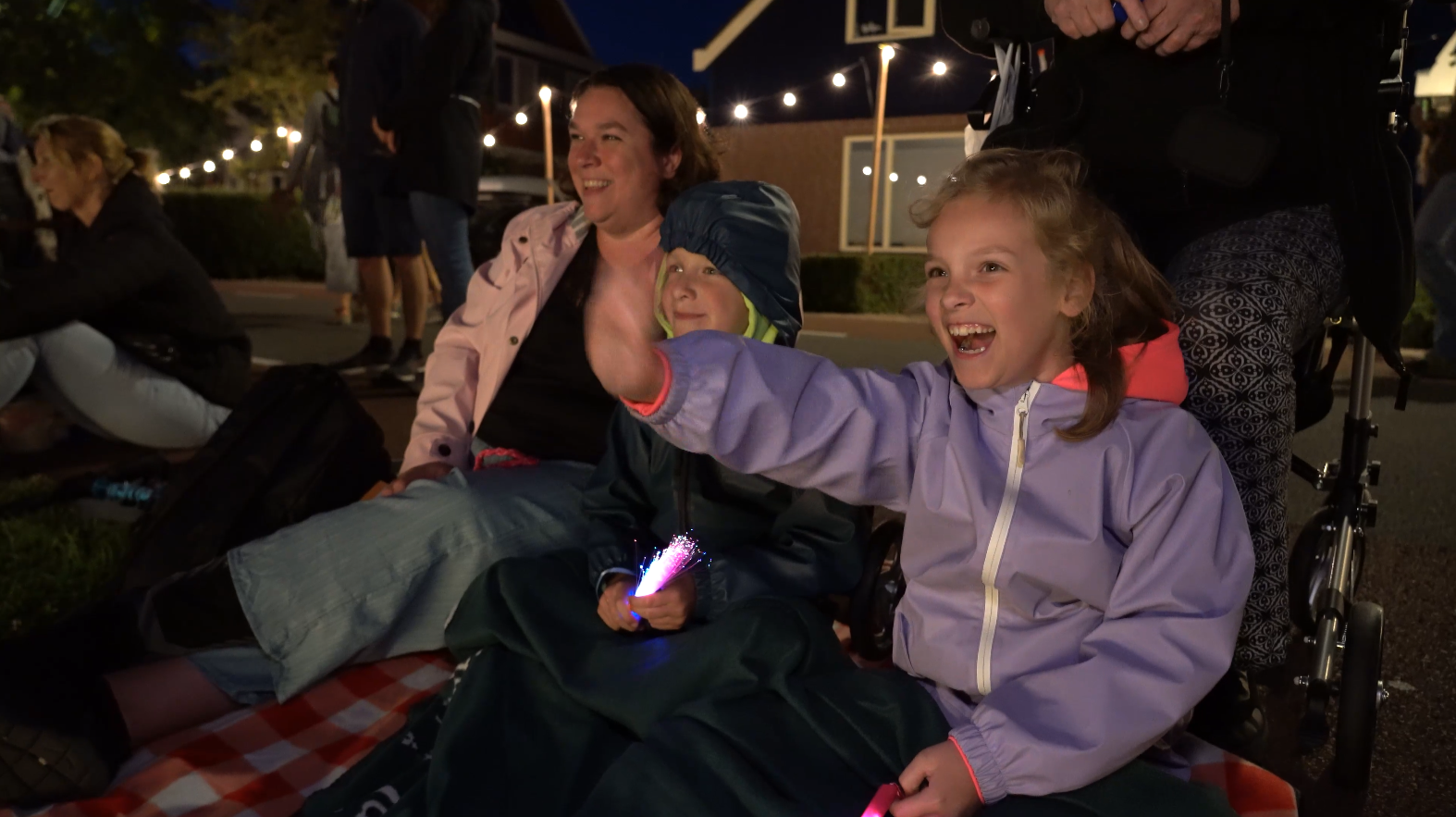 Een jonge meisje in een paarse jas lacht en zwaait enthousiast terwijl ze naast een vrouw en een jongen op een picknickkleed zit, omringd door andere mensen bij een avond evenement.