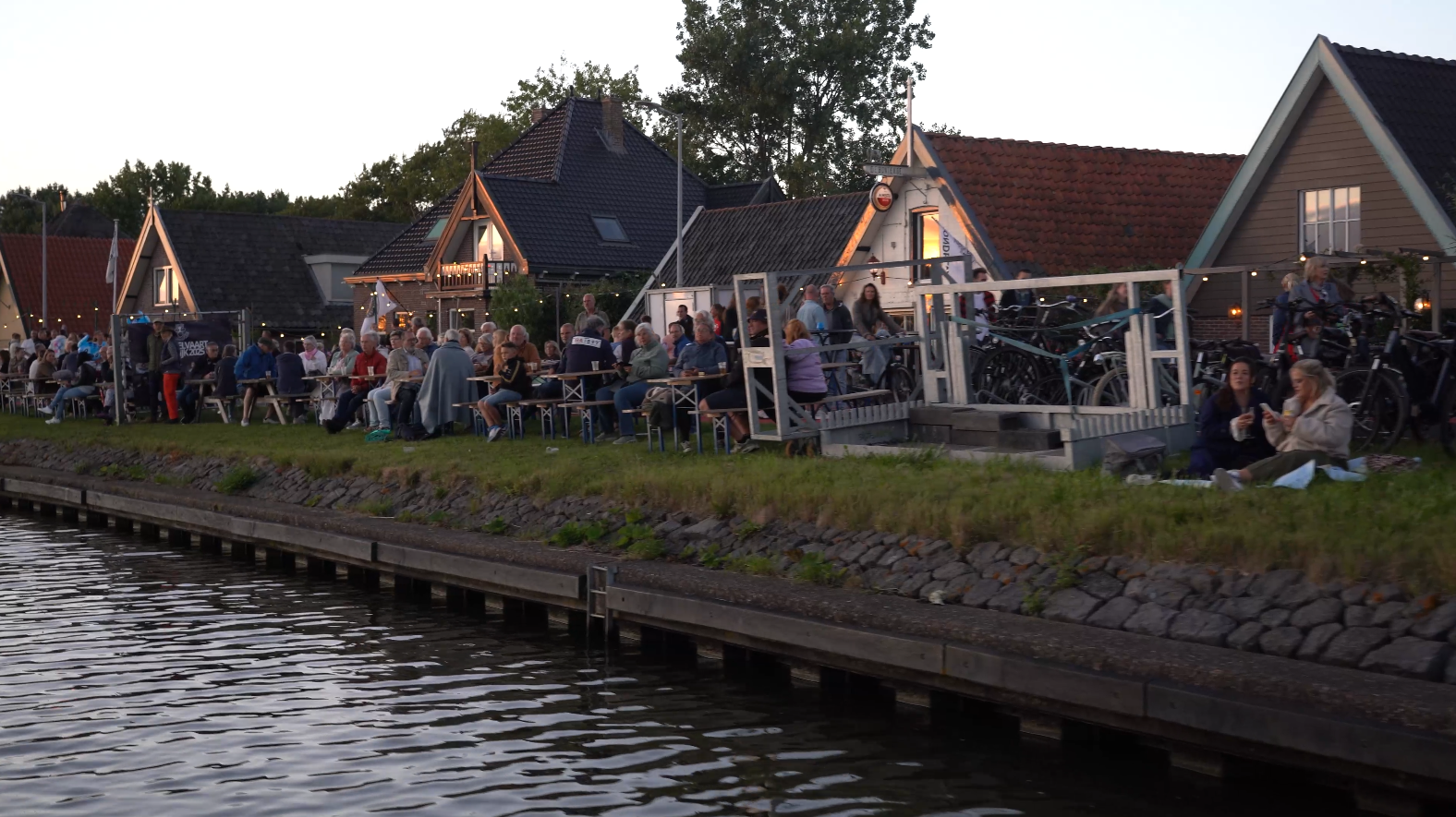 Groep mensen die 's avonds buiten zitten aan tafels langs een waterkant met huizen en lampjes op de achtergrond, enkele fietsen geparkeerd.