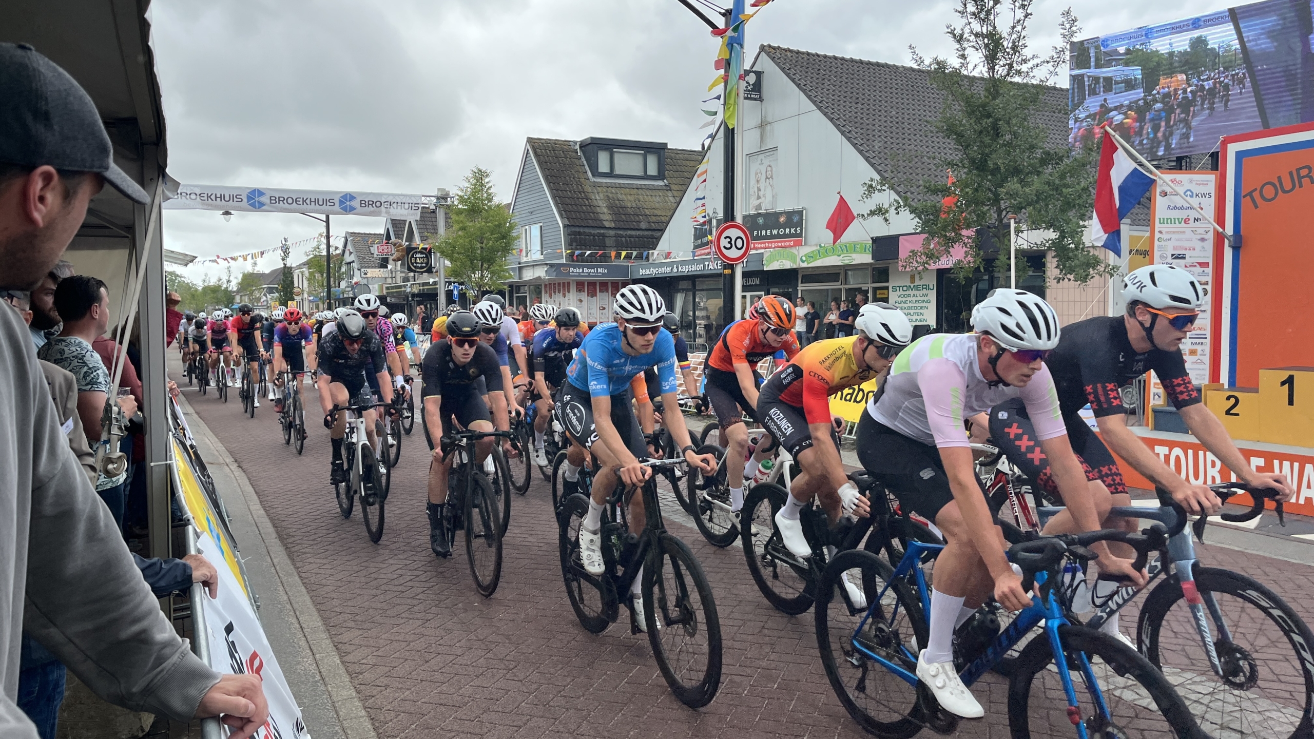 Groep wielrenners tijdens een wedstrijd in een stadsomgeving, omringd door toeschouwers en reclameborden.