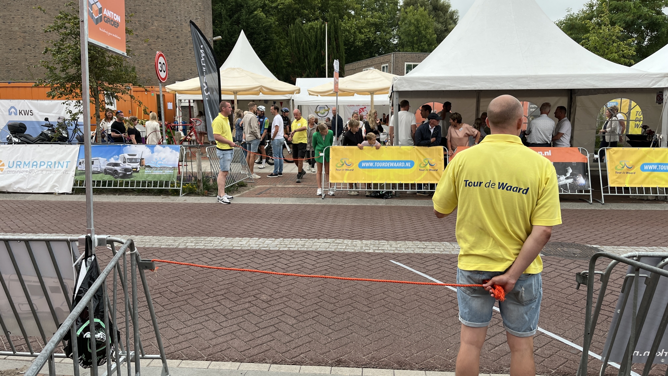 Een man in een geel "Tour de Waard" T-shirt staat voor een grensafzetting bij een evenement, met tenten en toeschouwers op de achtergrond.