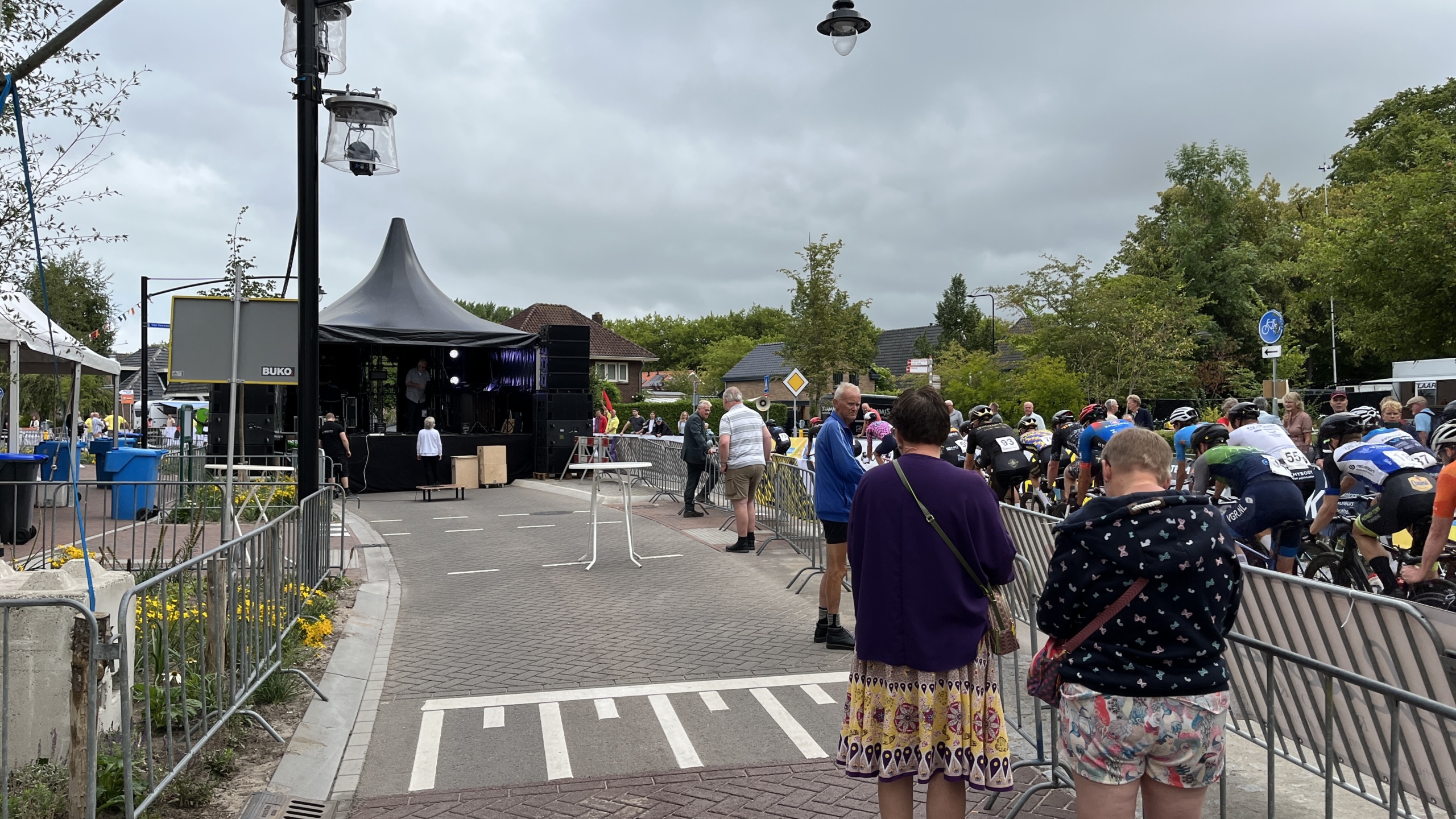 Een wielerwedstrijd met deelnemers die zich voorbereiden aan de startlijn, omringd door publiek en metalen barricades. Naast de straat, onder een zwart tentdak, staat een podium.