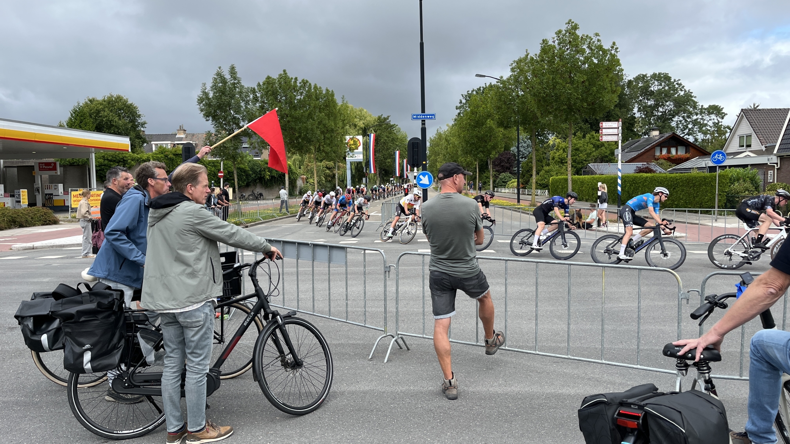 Wielrenners passeren een afzetting tijdens een race, terwijl toeschouwers en een man met een rode vlag langs de kant toekijken.