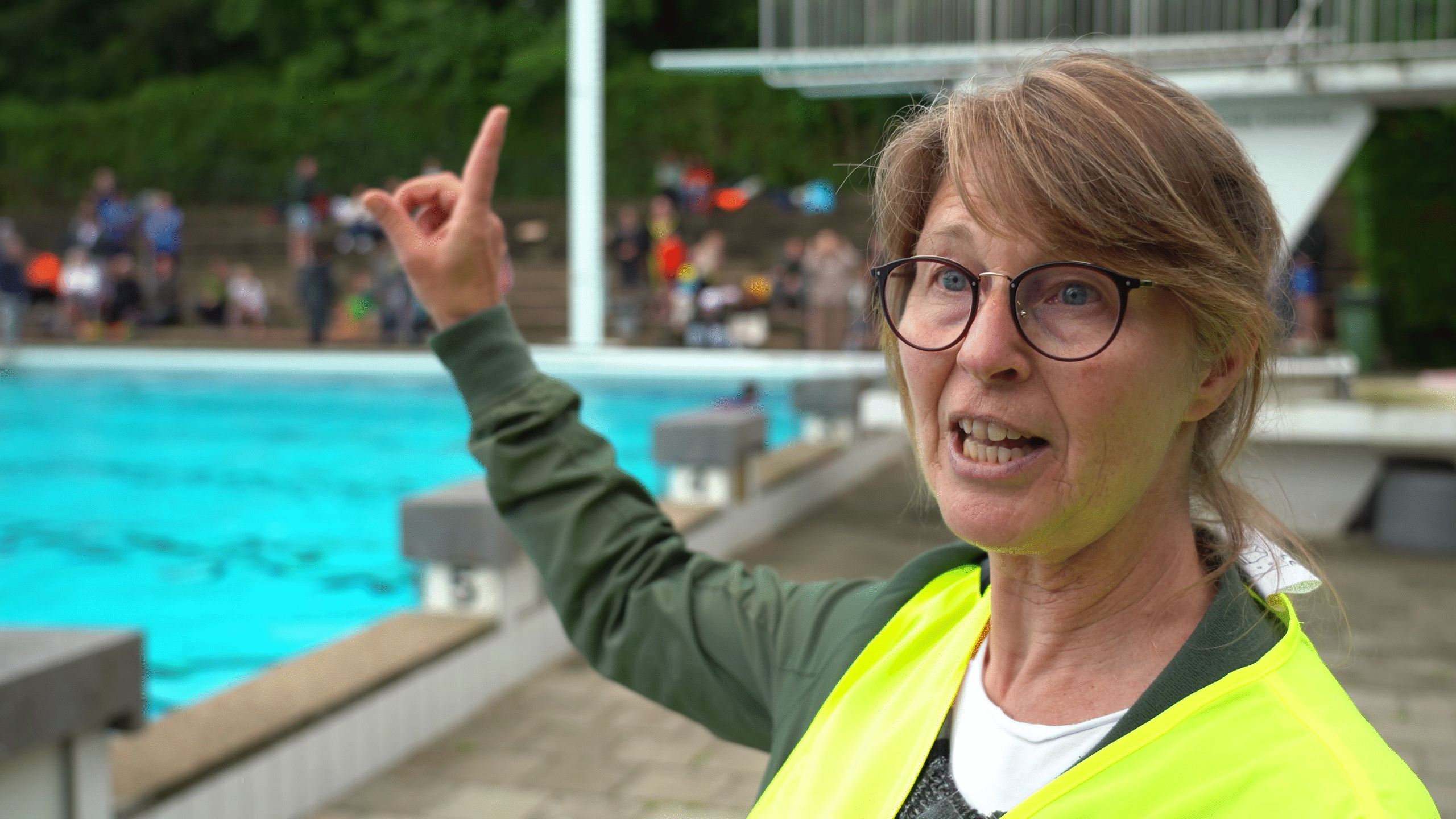 Een vrouw met bril en geel veiligheidsvest wijst met haar vinger naar iets in de verte bij een buitenzwembad, met mensen op de achtergrond.