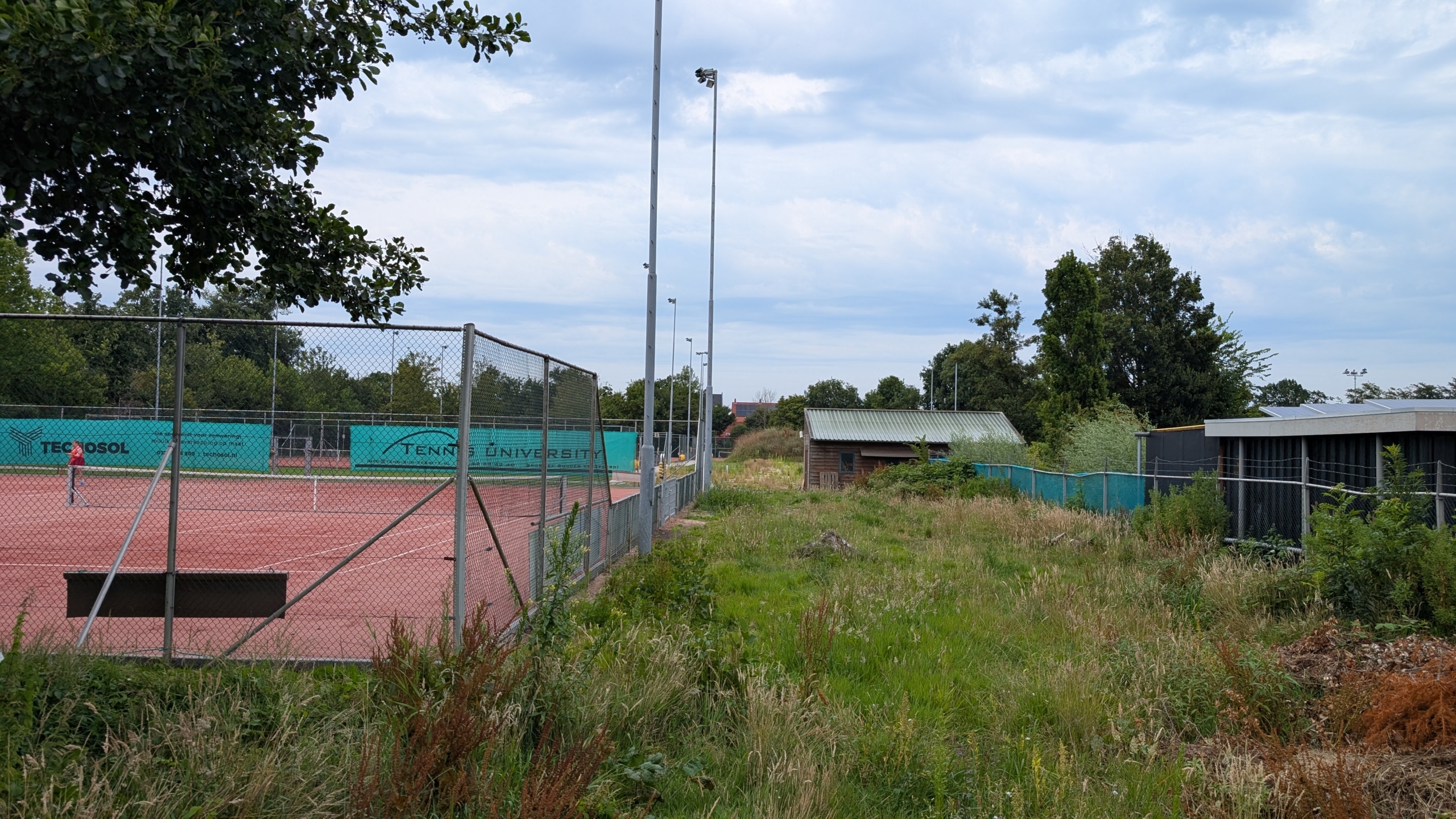 Een tenniscourt omheind met gaas ligt naast een stuk begroeit grasland met bomen in de achtergrond. Op de baan is iemand bezig met tennissen. Er zijn lichten boven de omheining en schuren aan de rechterkant. De lucht is bewolkt.