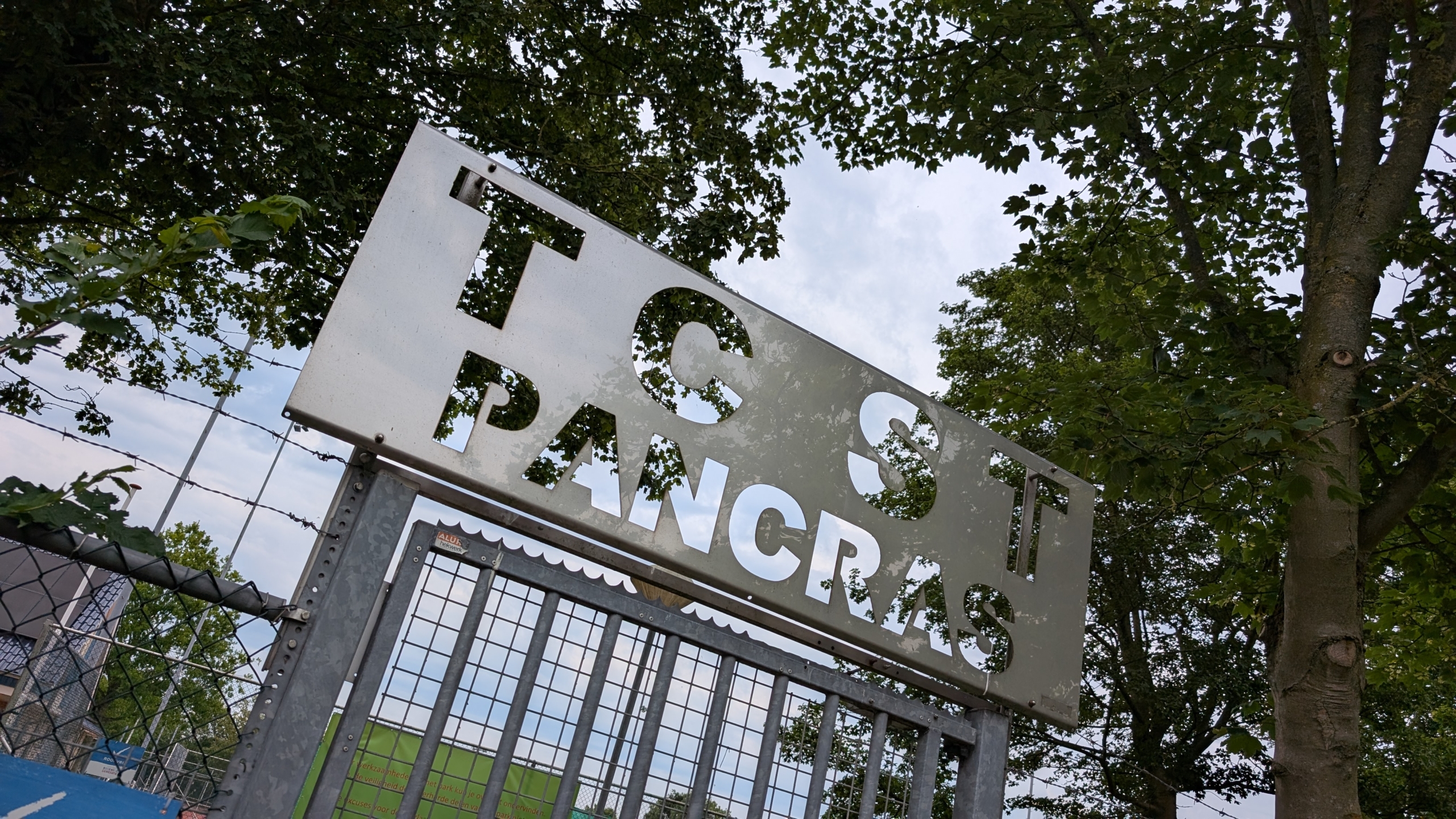 Een metalen bord boven een hek met de tekst "TC St Pancras", omgeven door bomen en takken.
