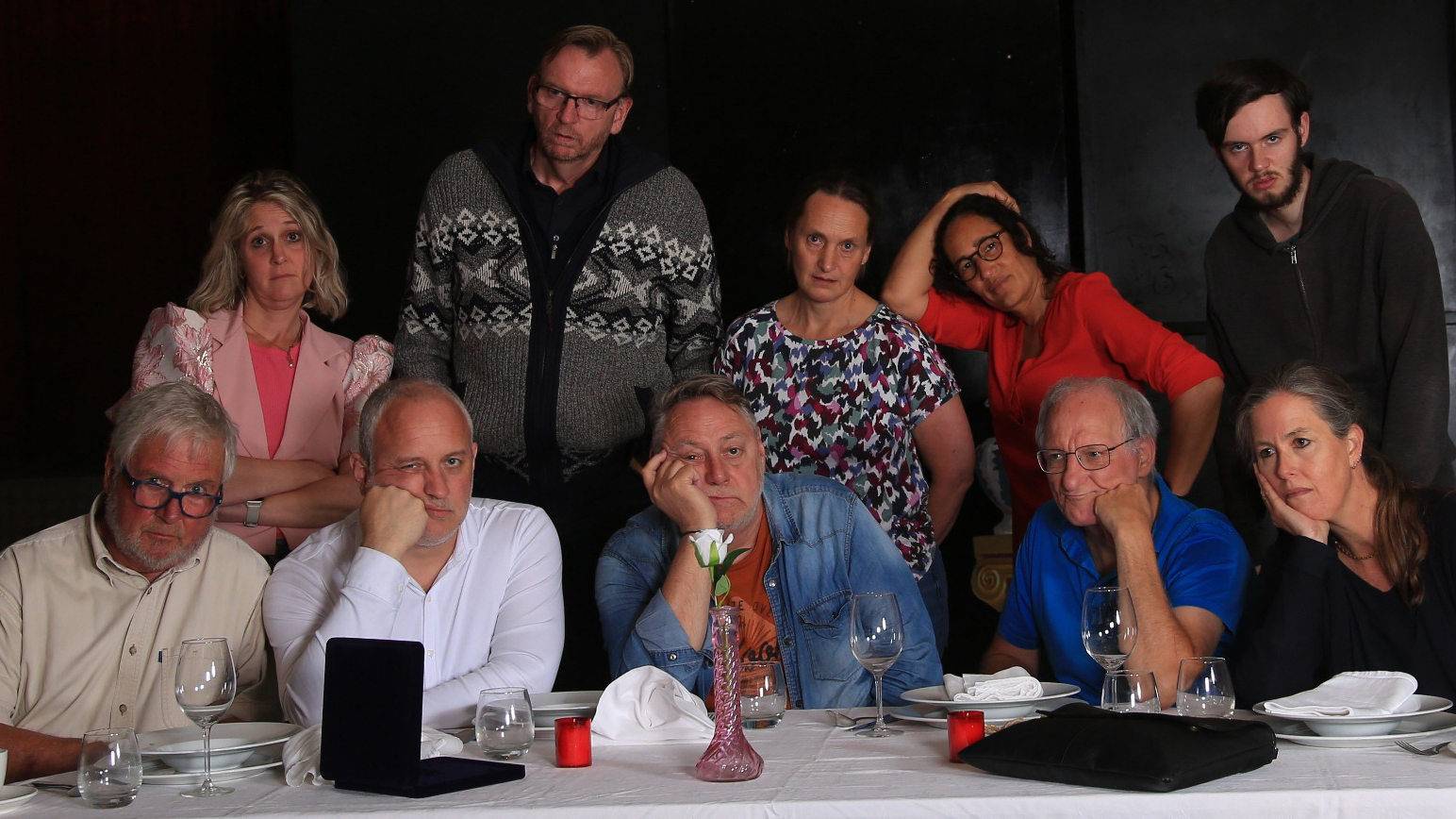 Groep van tien mensen staat en zit rond een tafel, kijken serieus en peinzend; de tafel is gedekt met glazen en borden.