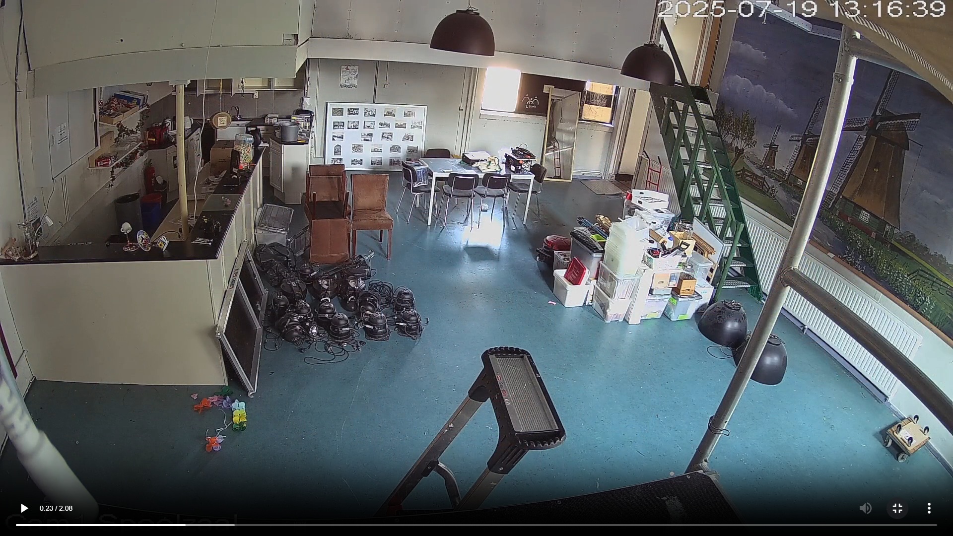 Een kamer met een blauwe vloer, een tafel, stoelen, en een stapel dozen. Aan de rechterkant is een schilderij van molens zichtbaar en er staat een ladder tegen de muur. Er liggen verschillende elektrische apparaten op de grond.