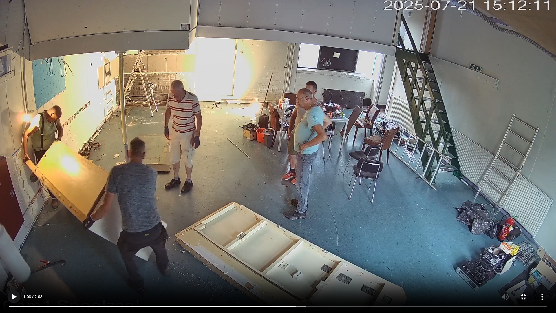 Mensen in een grote kamer zijn bezig met een verbouwingsproject, omringd door gereedschap en ladders. Enkele personen lijken een houten paneel te verplaatsen, terwijl anderen toekijken. De kamer heeft een blauwe vloer en een metalen trap aan de rechterkant.