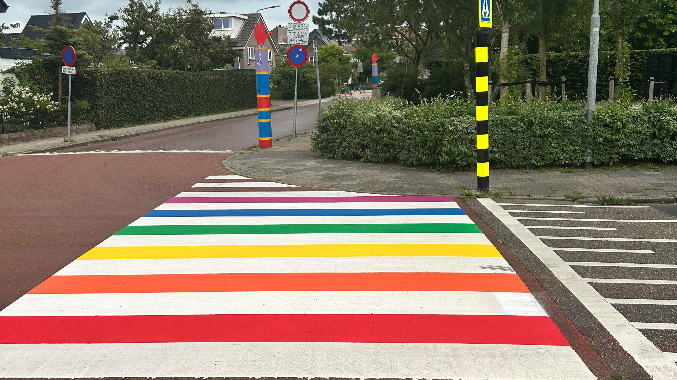 Een zebrapad in regenboogkleuren op een straat met verkeersborden en paaltjes ernaast.