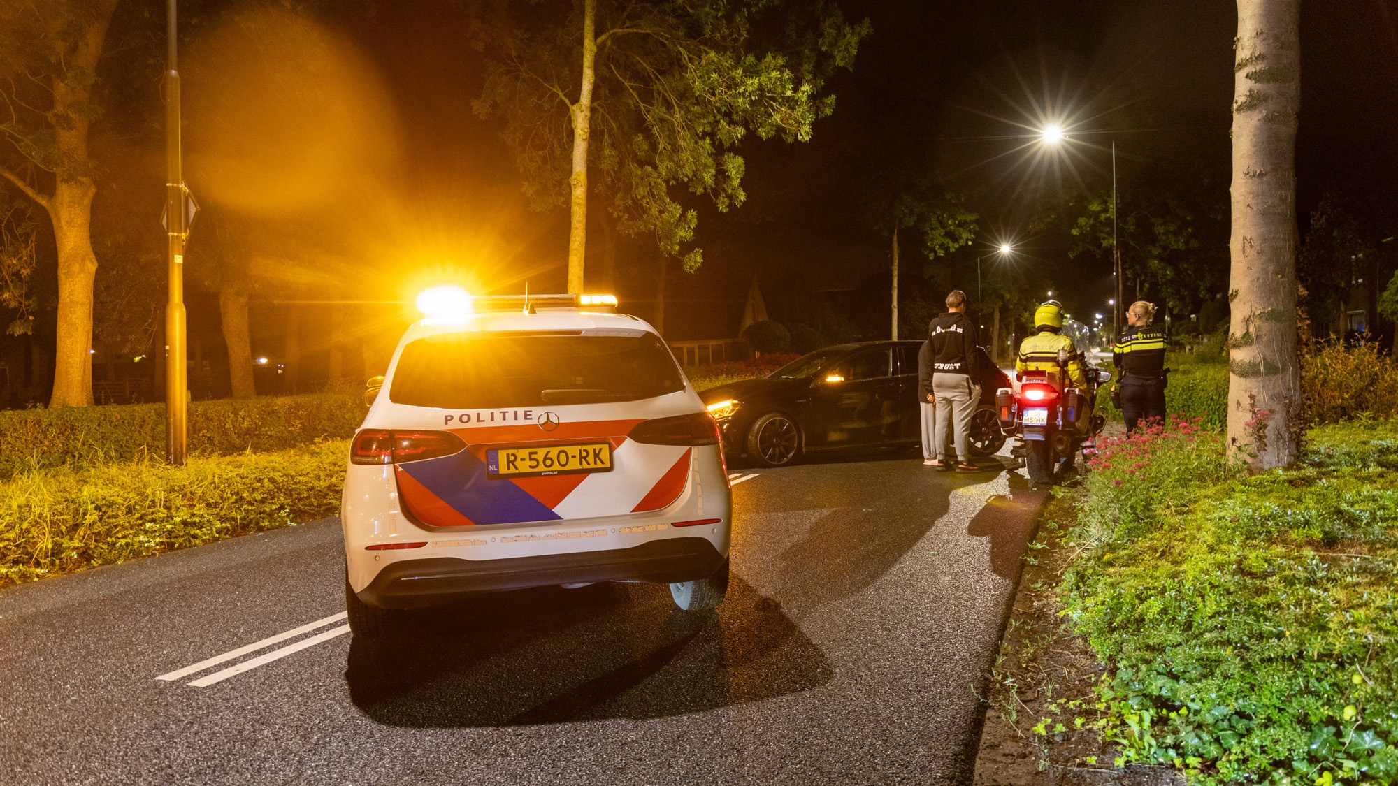 Een politieauto met draaiende lichten en motorrijders staan 's nachts langs een weg met een stilstaande auto, terwijl twee mensen met een politieagent praten.