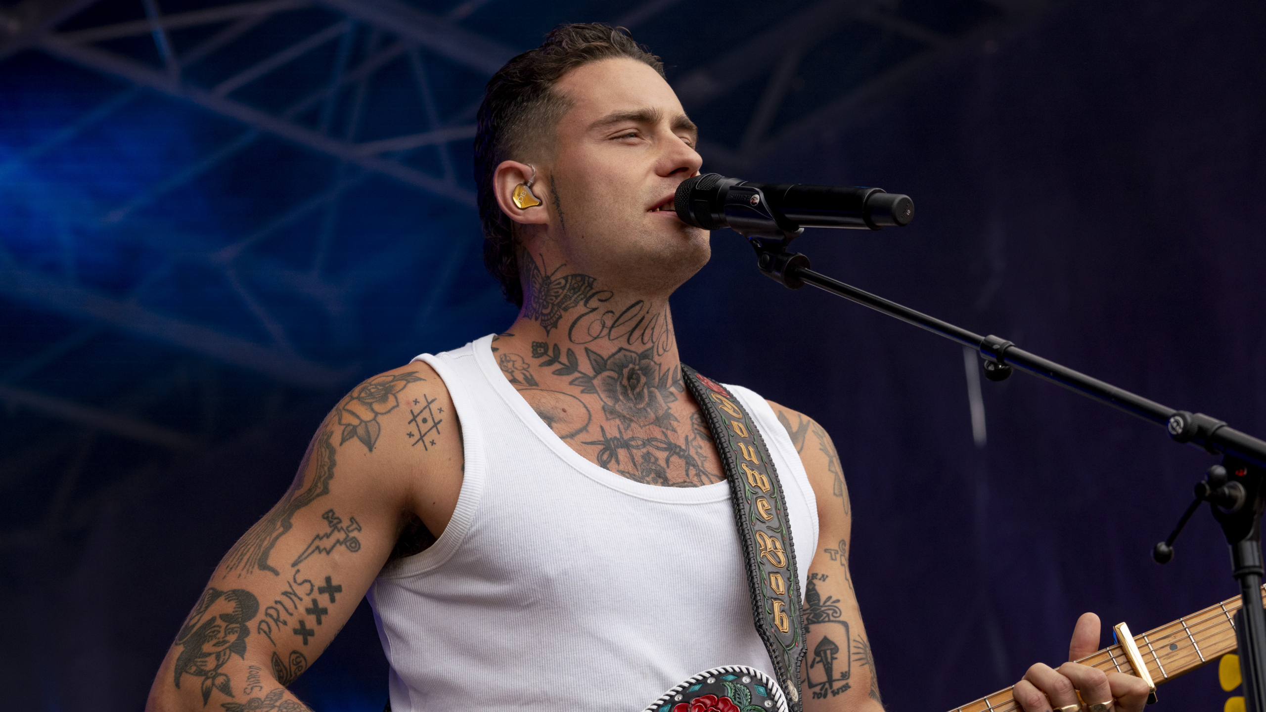 Man met tattoos die in een microfoon zingt tijdens een optreden, terwijl hij een gitaar speelt. Hij draagt een wit hemd en gele oordopjes.