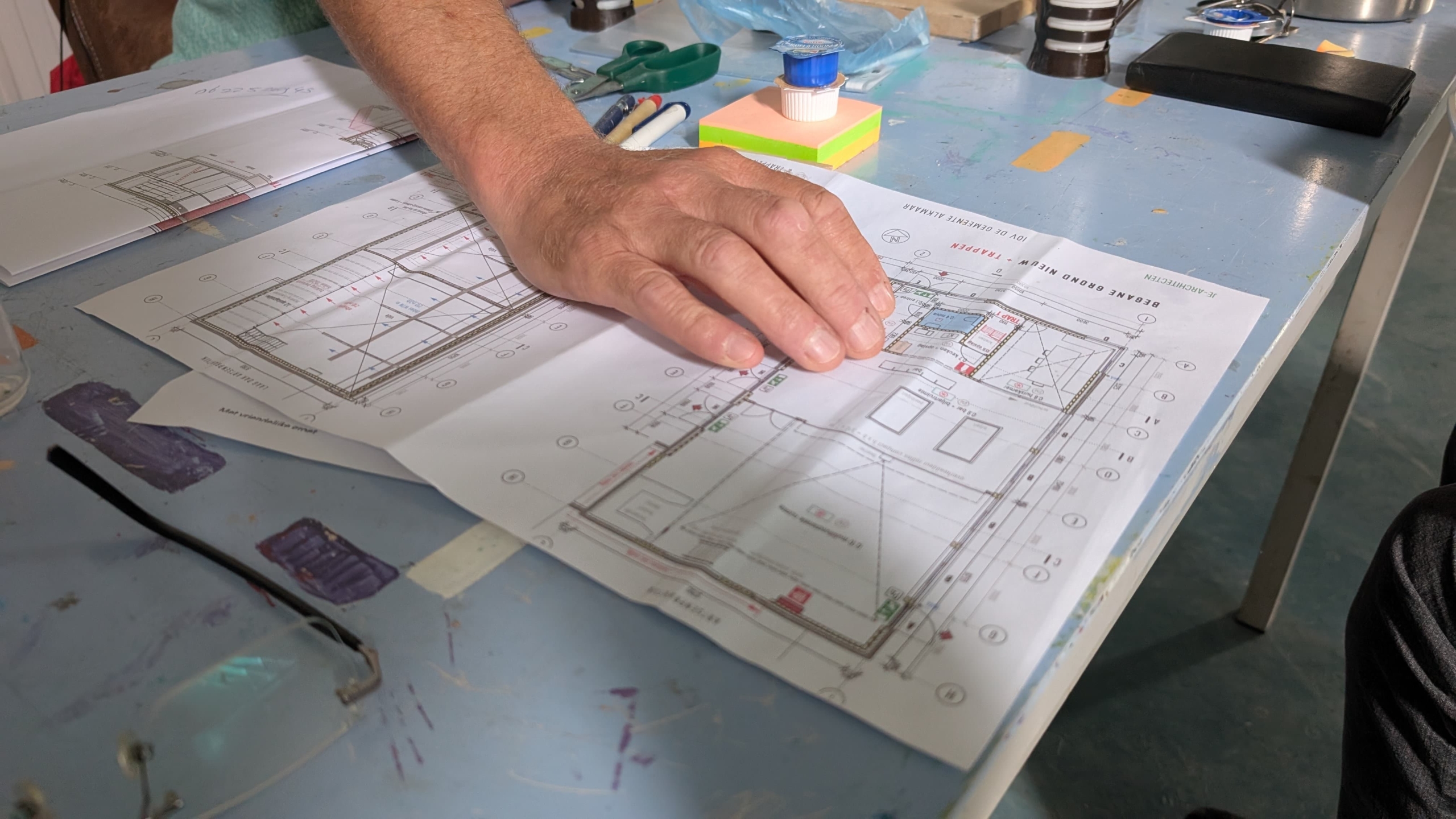 Een hand rust op een gedetailleerde bouwtekening, omringd door tekenspullen, op een tafel met een blauwe verflaag.
