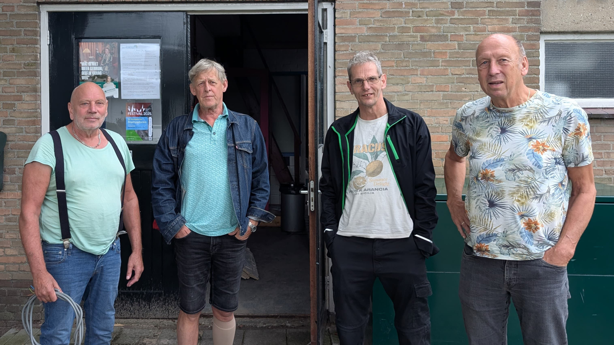 Vier mannen staan buiten een gebouw, waarvan de deur openstaat, met posters erop. Ze dragen casual kleding en lijken ontspannen. De achtergrond is een bakstenen muur met een klein raam.
