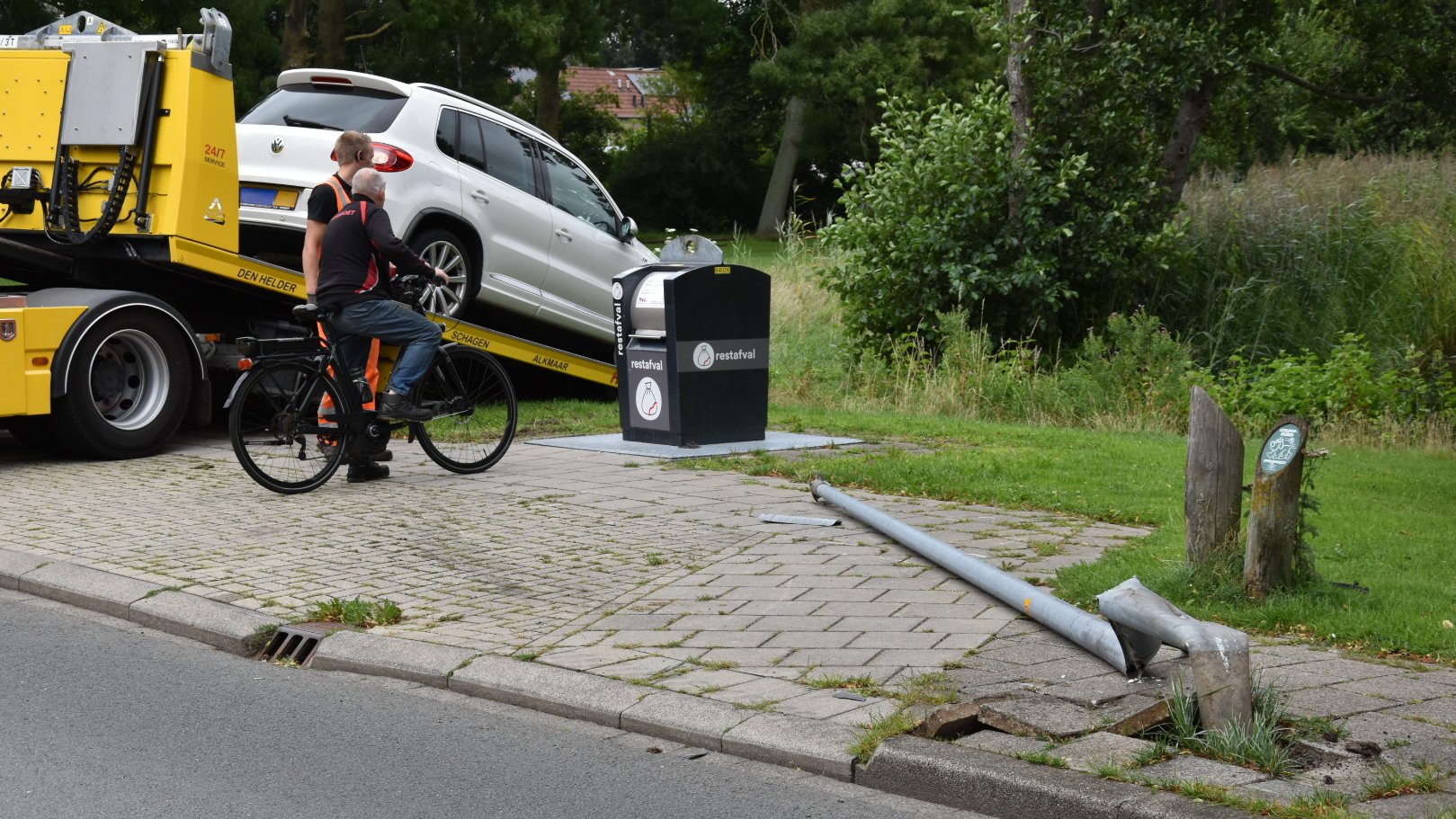 Een witte auto wordt weggesleept door een gele takelwagen, terwijl een lantaarnpaal omgevallen ligt op het trottoir. Een man en een vrouw op een fiets kijken naar de situatie. Op de achtergrond staan groene bomen en gras.
