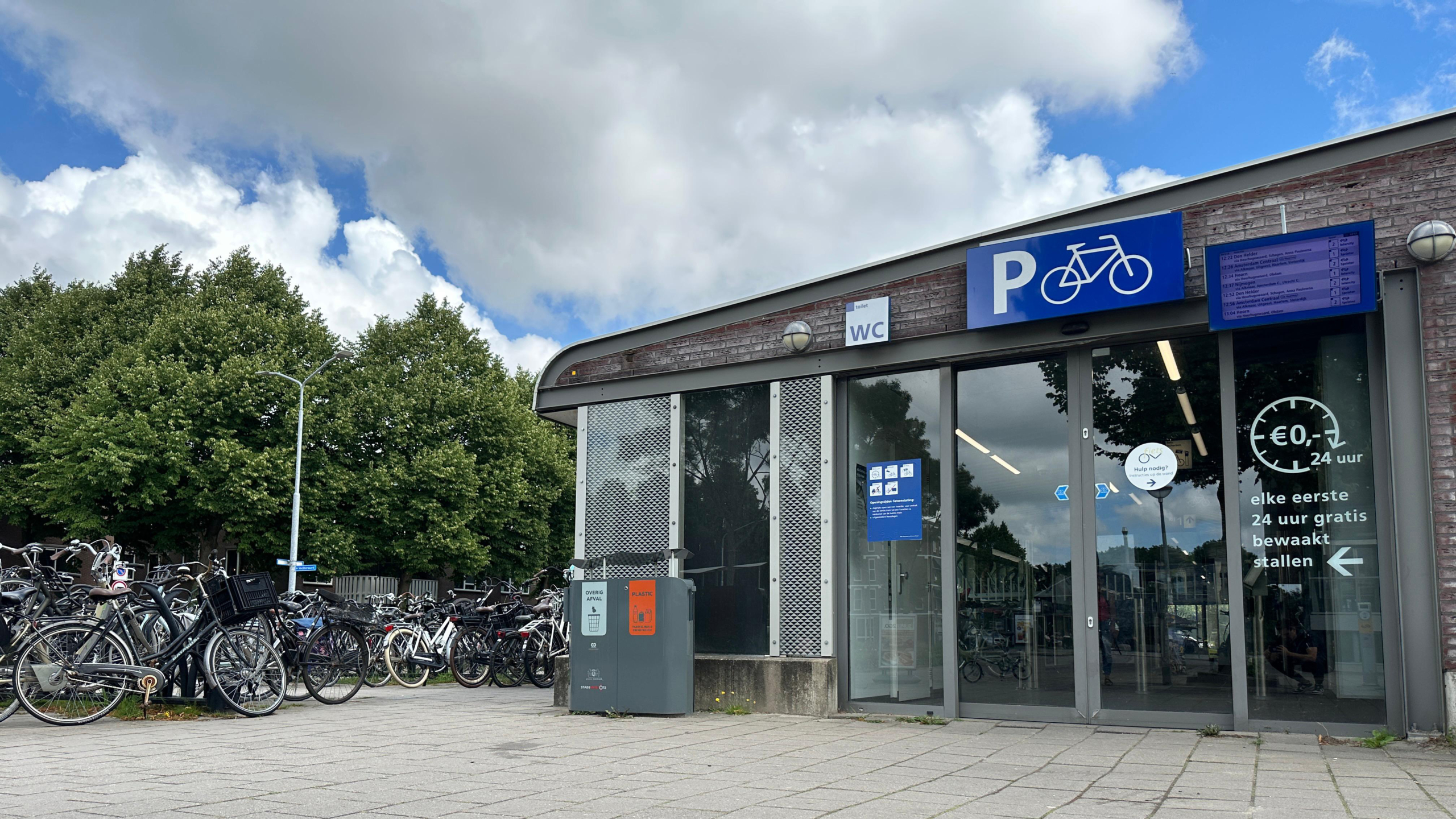 Fietsenstalling met meerdere fietsen geparkeerd naast een gebouw met glazen deuren, waarop een bord met een witte fiets op een blauwe achtergrond hangt en prijsaanduidingen te zien zijn. Er staan ook afvalbakken naast het gebouw en op de achtergrond zijn groene bomen en bewolkte lucht zichtbaar.