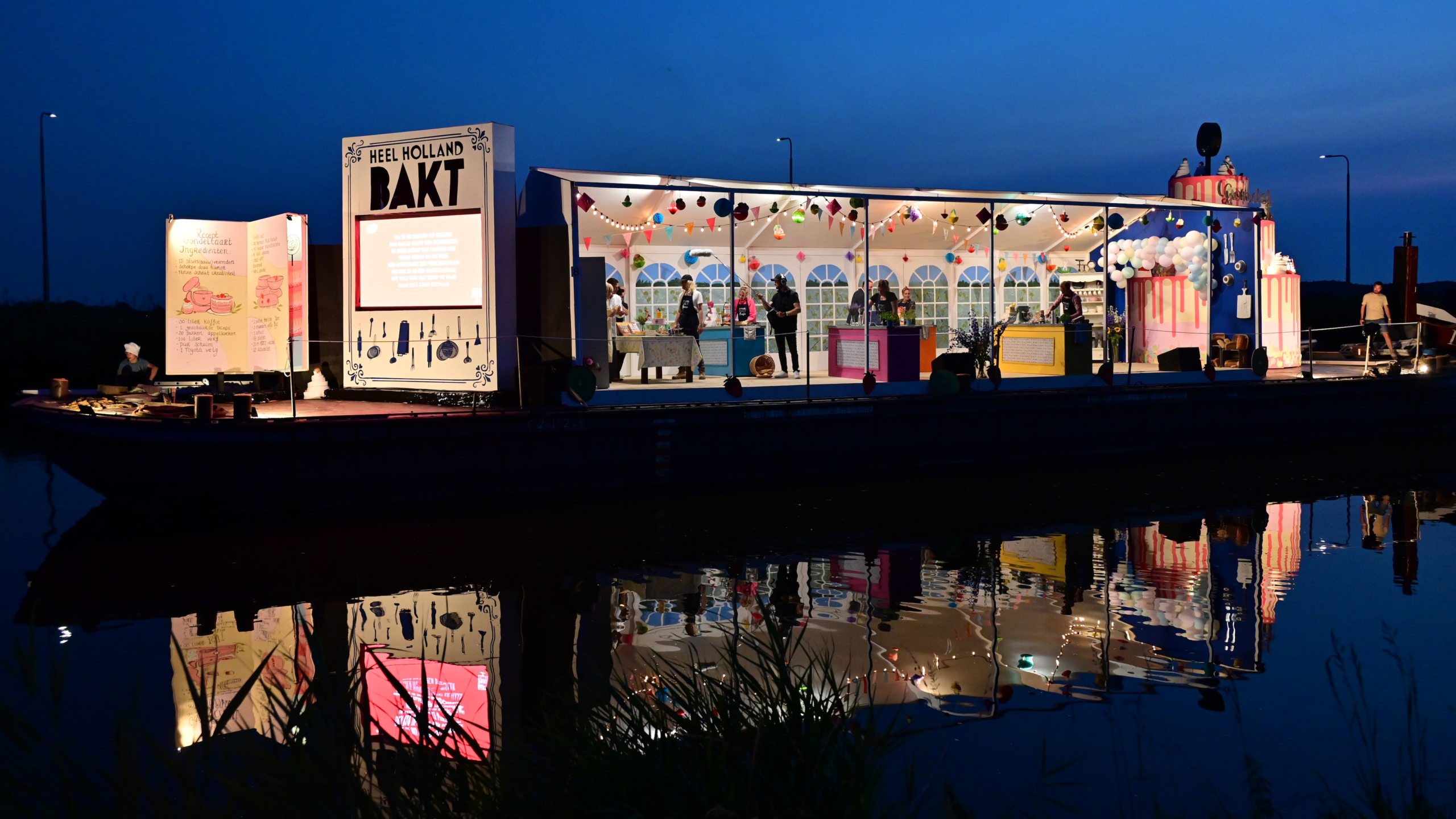 Een verlichte opstelling van "Heel Holland Bakt" aan het water in de avond; het heeft een open keuken met mensen binnen, versierd met lichtjes en kleurrijke decoraties.