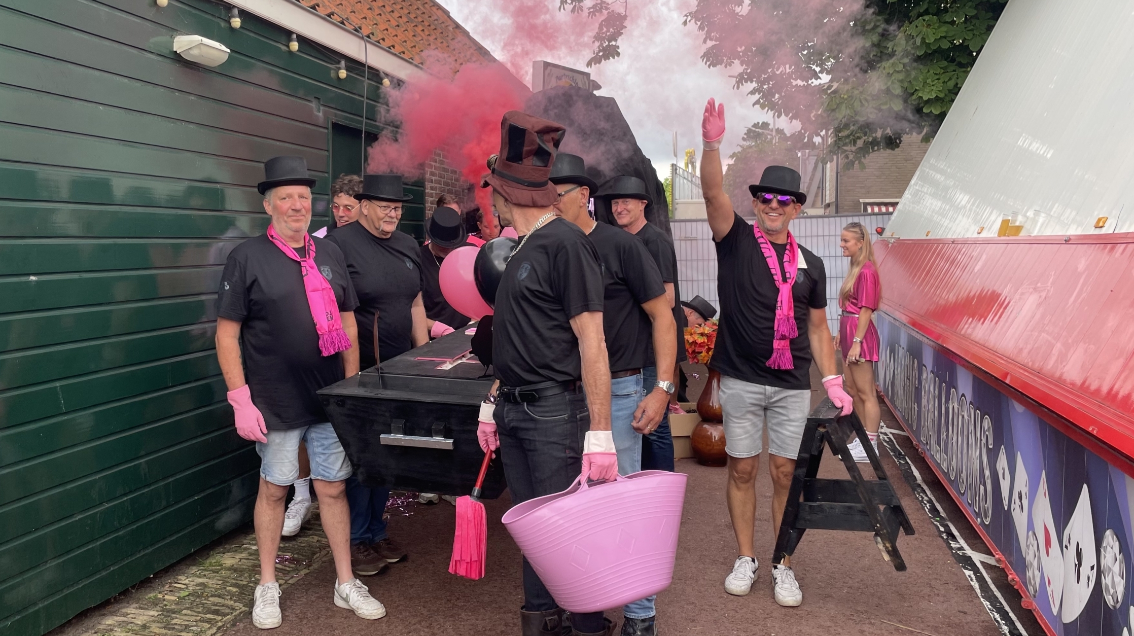 Een groep mannen in zwarte shirts en hoge hoeden staan buiten, sommige dragen roze accessoires zoals handschoenen en sjaals. Ze staan naast een zwarte kist en er hangt roze rook in de lucht. Achter hen is een groen gebouw. Eén man houdt een roze emmer en een ander zwaait met de hand omhoog. Rechts is een vrouw in een roze outfit te zien.