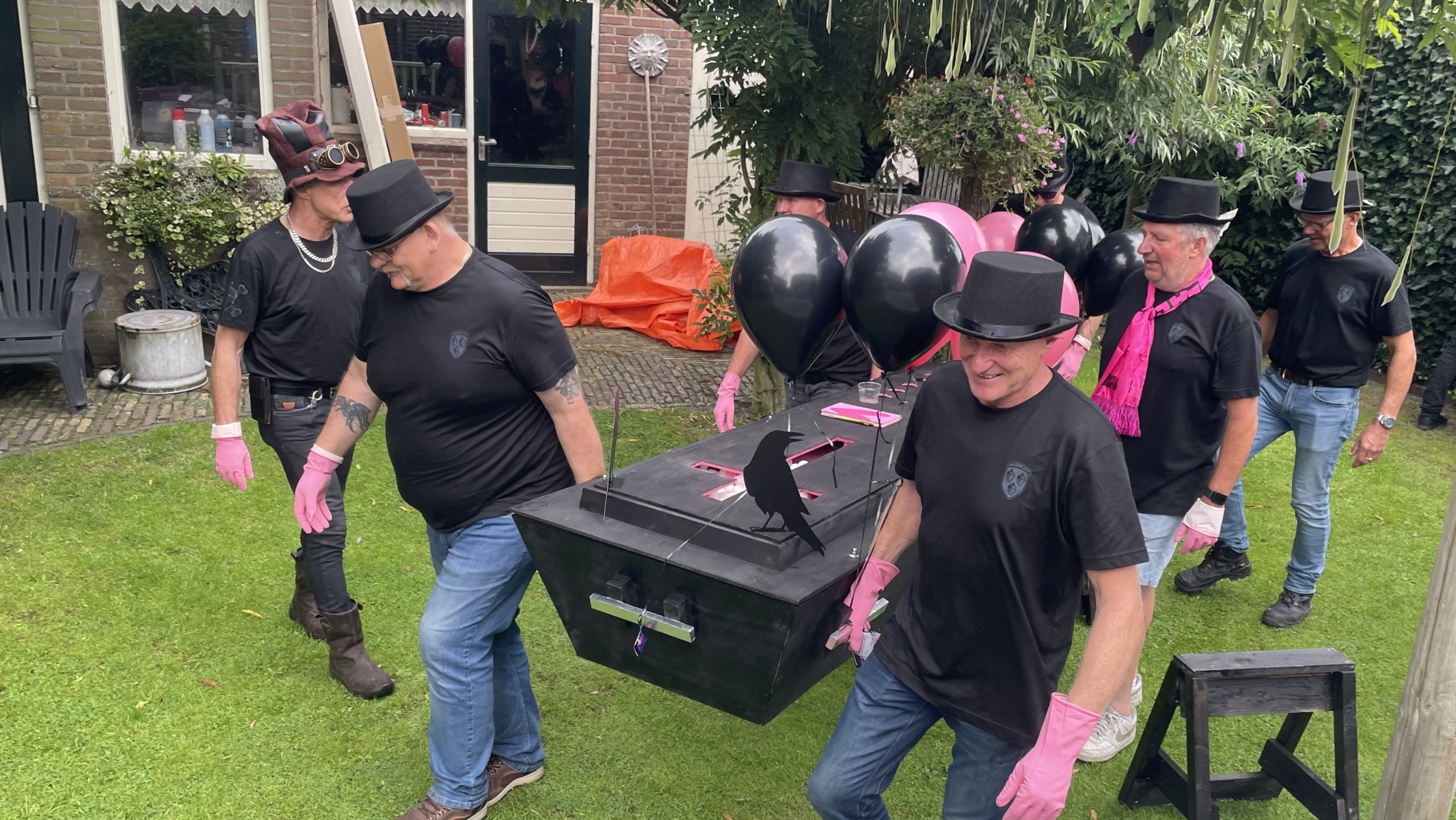 Een groep mannen gekleed in zwarte shirts en hoeden draagt een zwarte kist versierd met zwarte en roze ballonnen door een tuin. Ze dragen roze handschoenen, en een van hen draagt een roze sjaal. De tuin is omgeven door groen met een stenen huis op de achtergrond.