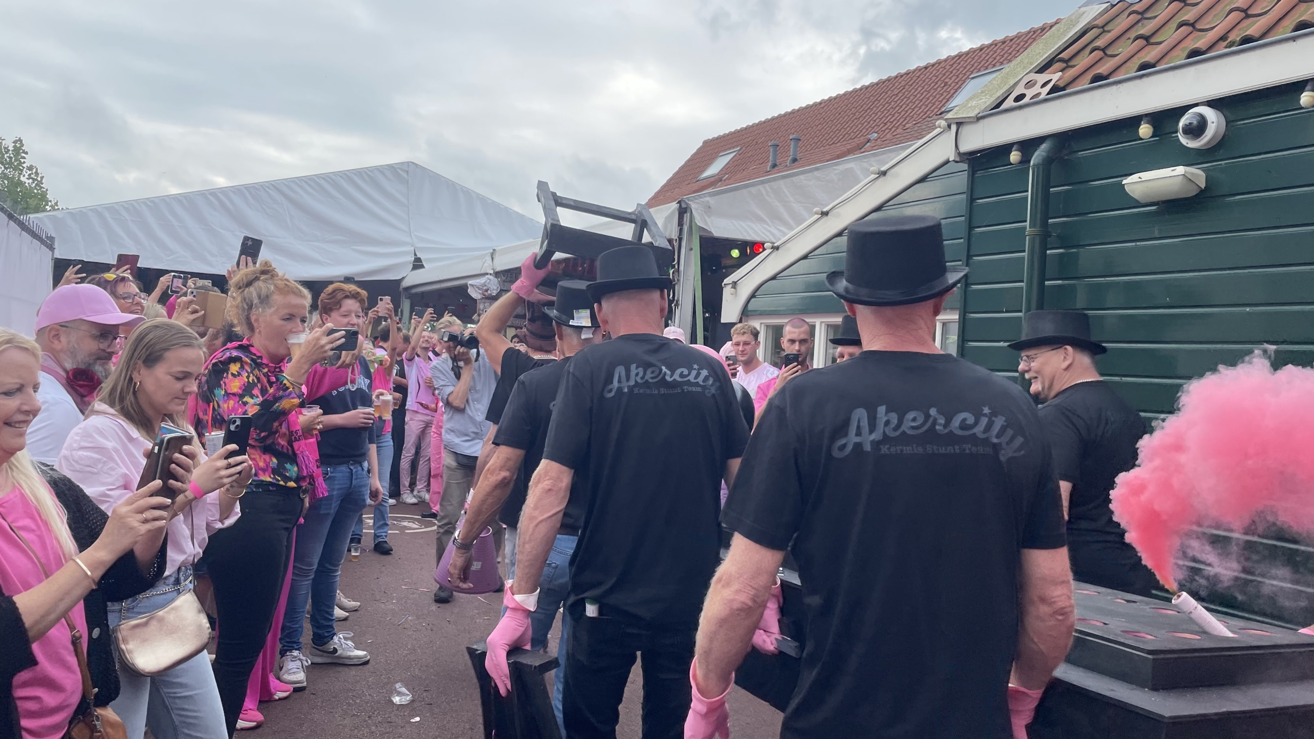 Groep mensen bij een evenement, met sommigen in zwarte hoeden en t-shirts met 'Akercity', terwijl een persoon iets omhoog tilt. Omstanders maken foto's en een roze rookwolk is te zien.