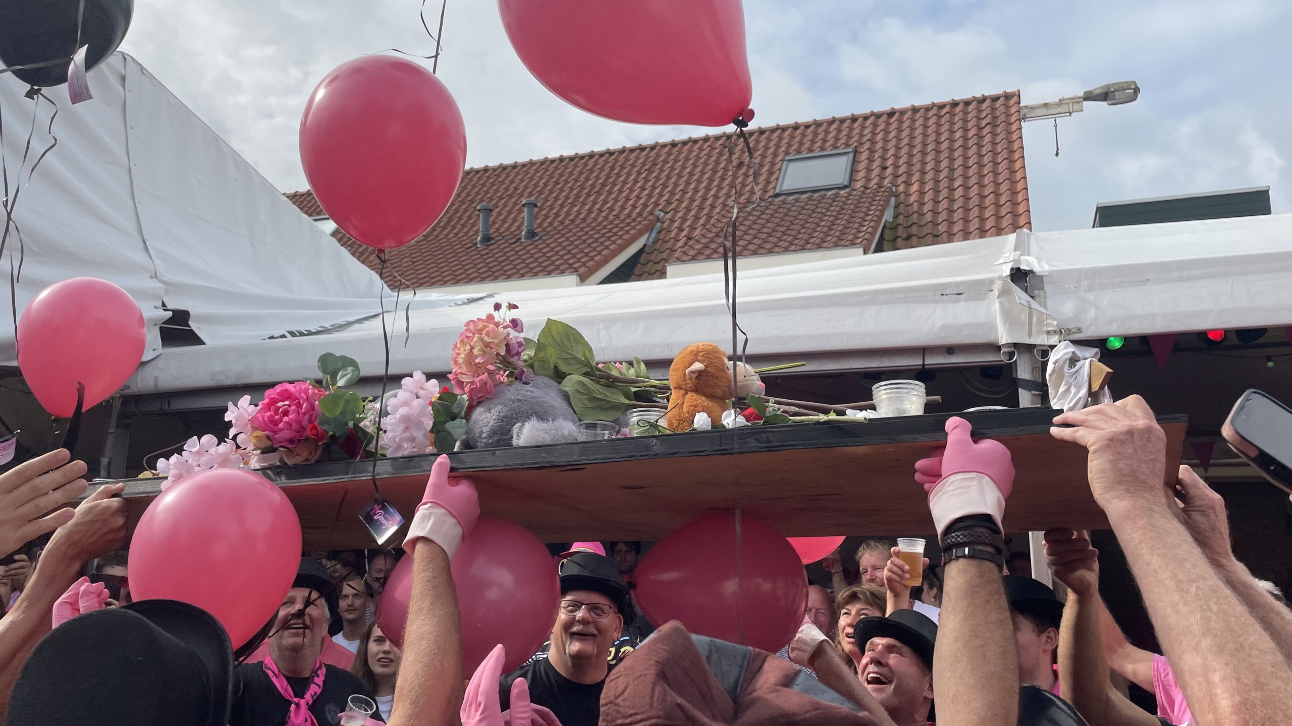 Groep mensen verzameld onder een houten plank versierd met bloemen, ballonnen en knuffels, opgehouden door personen met roze handschoenen.