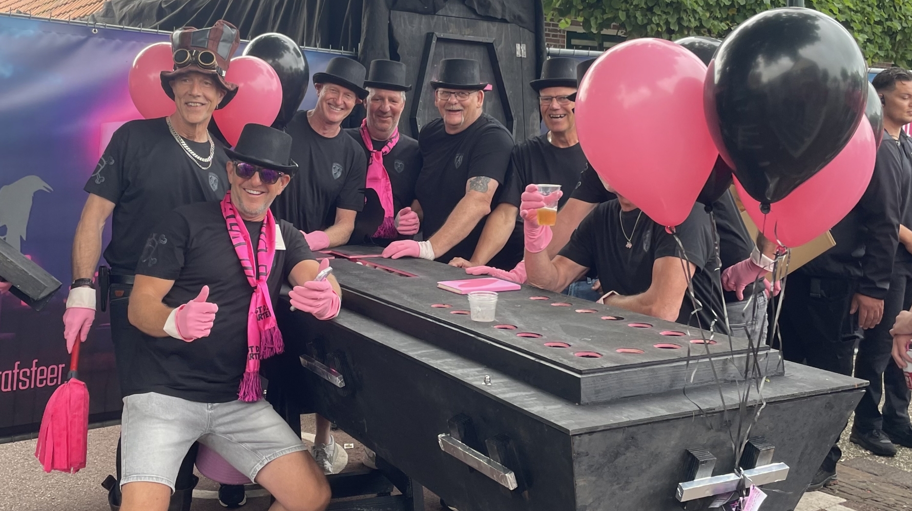 Een groep mannen in zwarte kleding en hoedjes poseert lachend rondom een zwart, doodskist-vormig voorwerp. Ze dragen roze handschoenen en sjaals, en er zijn roze en zwarte ballonnen te zien. Op de achtergrond is een gebouw met een rood dak en een reclamebord zichtbaar.