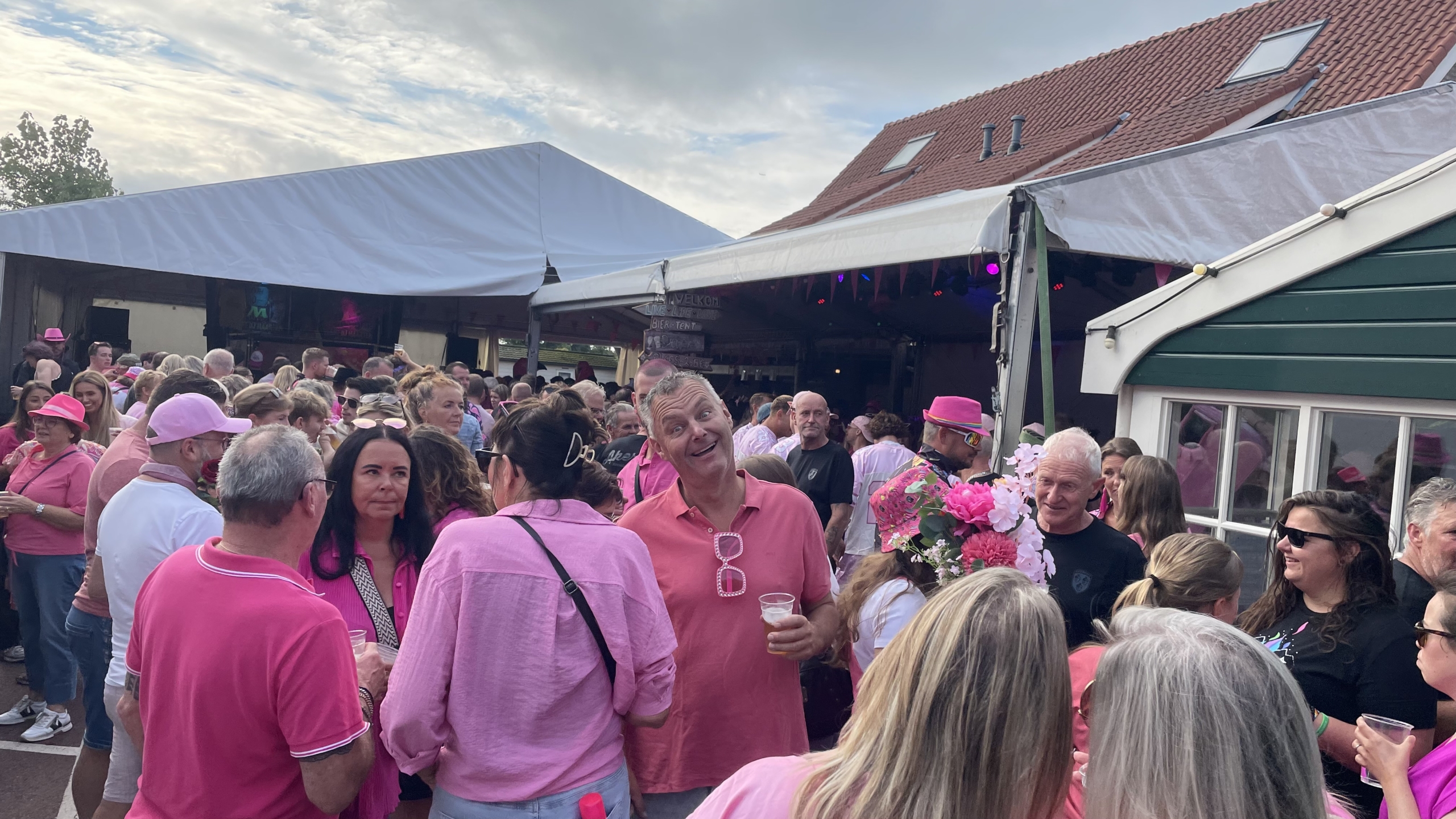 Mensenmassa bij een buitenevenement, voornamelijk gekleed in roze, met feesttenten en tenten op de achtergrond.