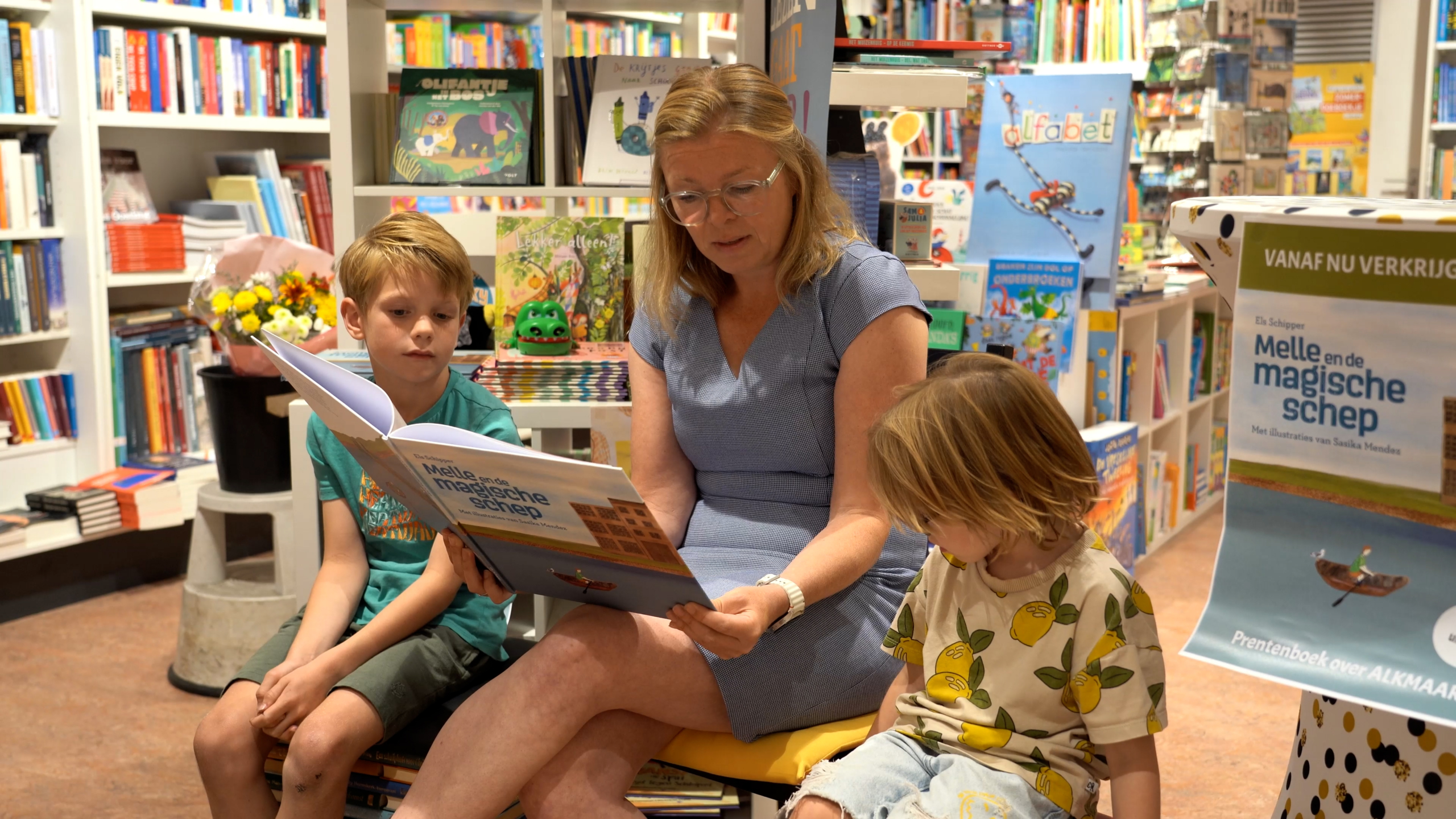 Een vrouw leest voor aan twee kinderen in een boekwinkel, omringd door planken met boeken en kinderliteratuur.