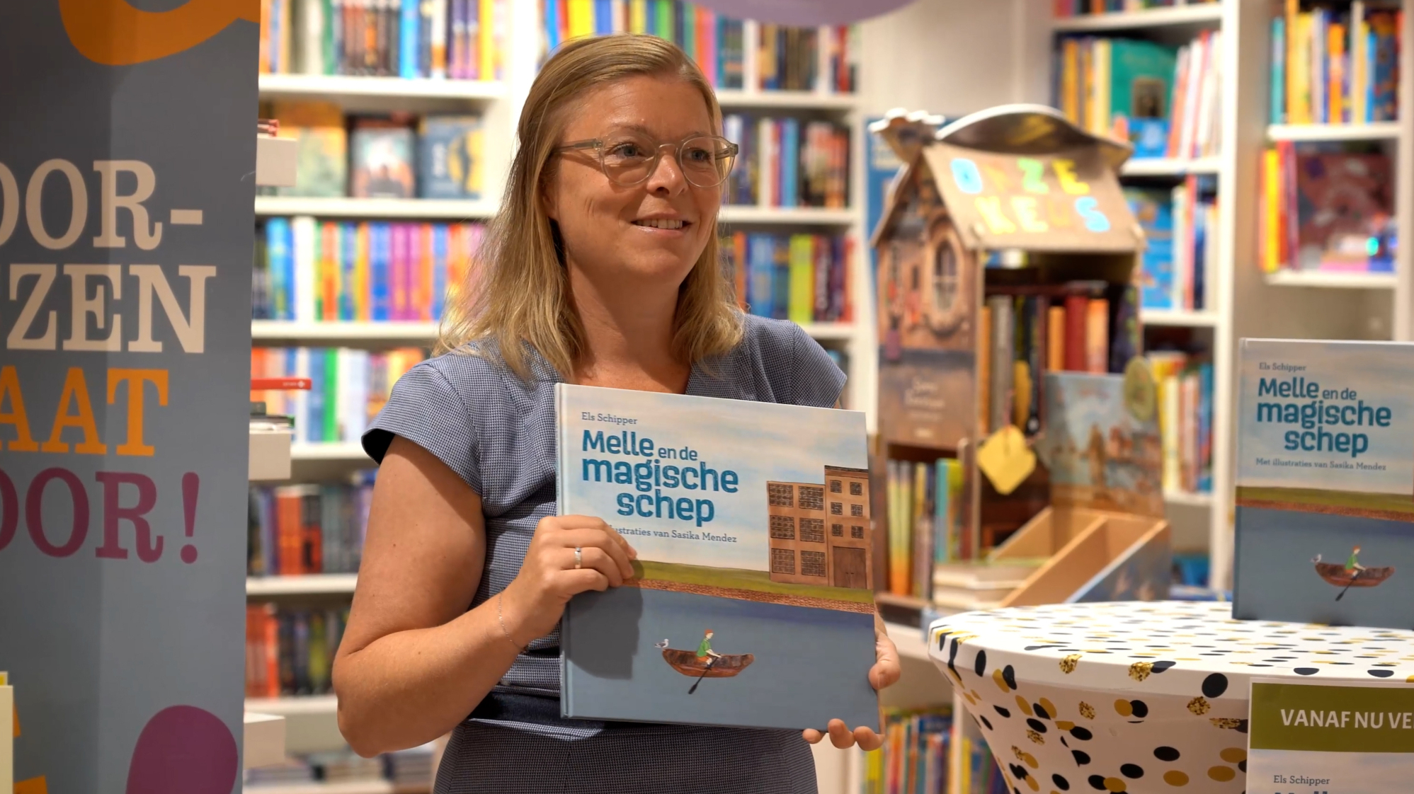 Een vrouw houdt het kinderboek 'Melle en de magische schep' vast in een boekwinkel, omringd door kleurrijke boekenplanken; op de tafel ligt een exemplaar van hetzelfde boek.