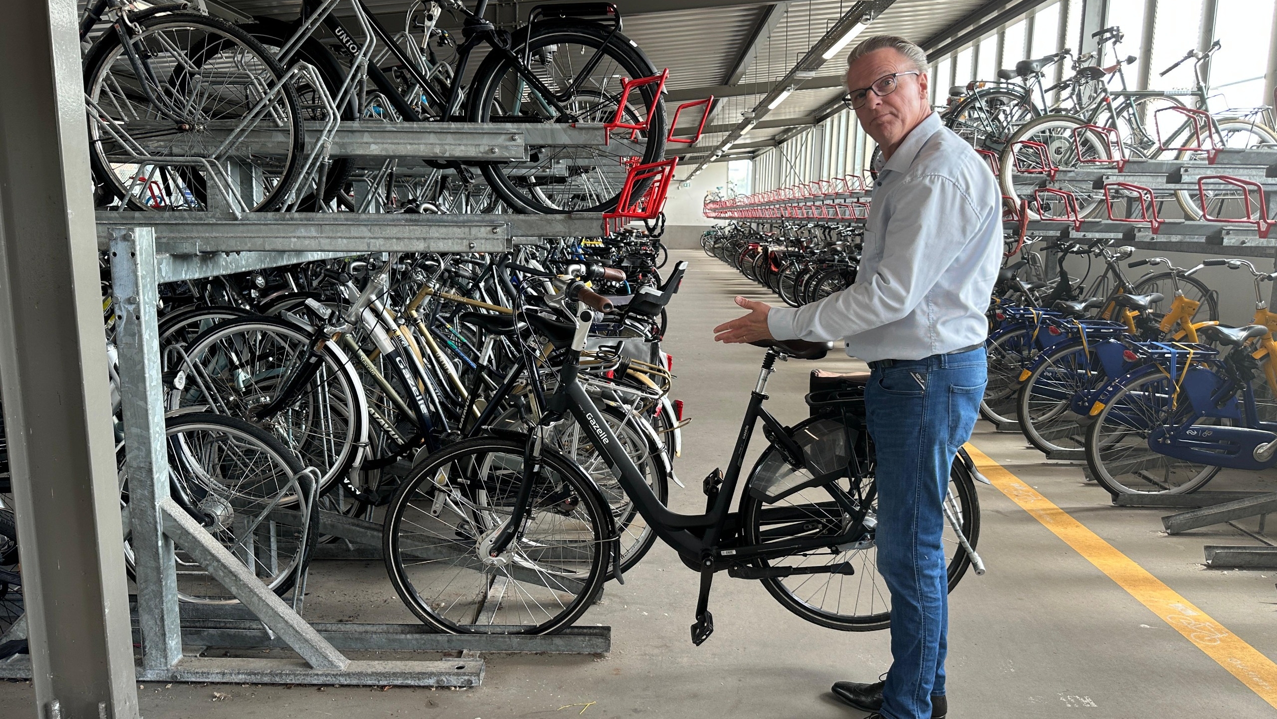 Man in casual clothing standing in a large bicycle storage area with multiple bikes parked on two levels of racks.