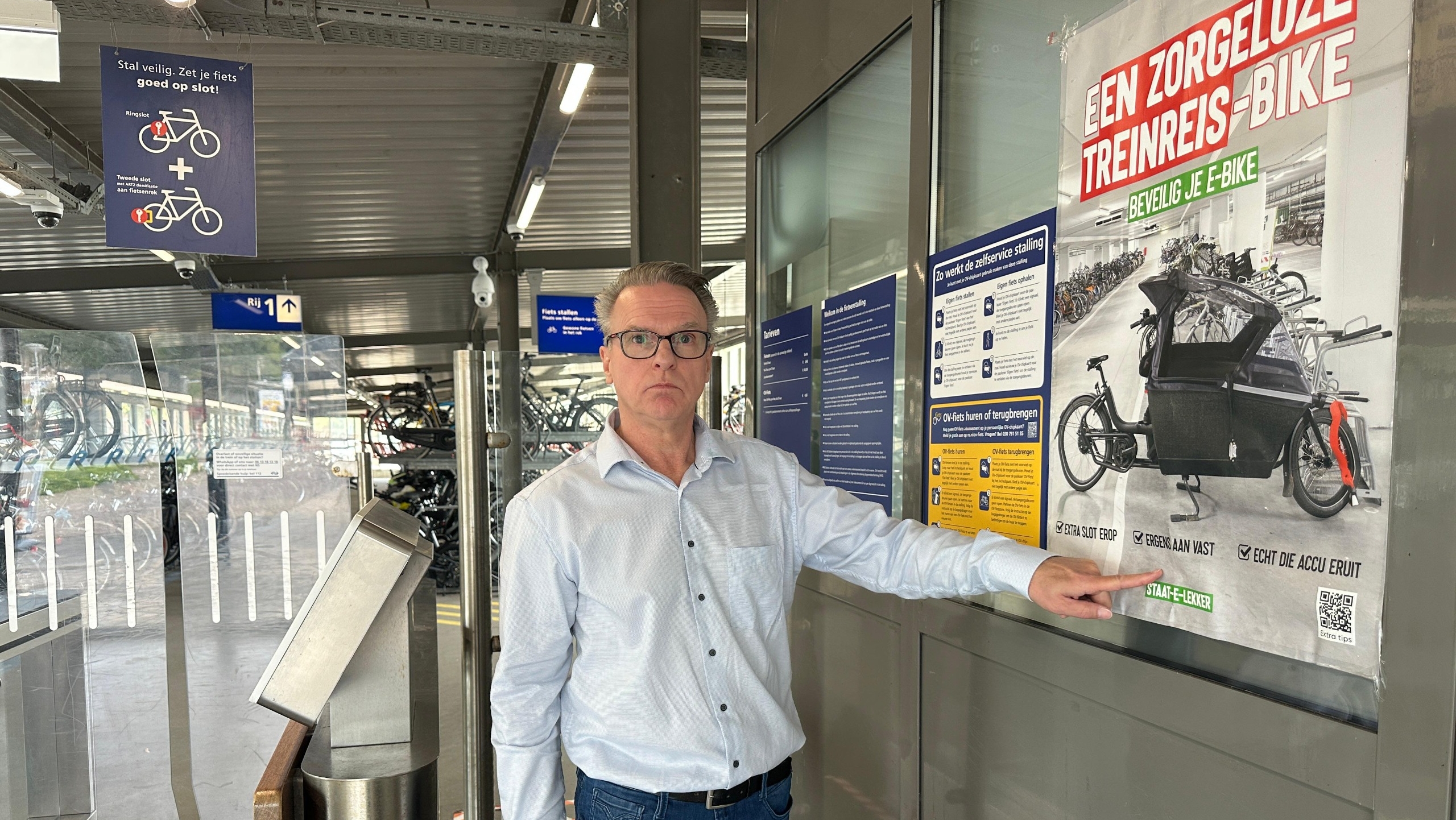 Een man in een blauwe overhemd staat in een fietsenstalling naast een poster die tips geeft voor het veilig stallen van e-bikes. De poster benadrukt het belang van het vastzetten van de fiets en het verwijderen van de accu. De omgeving bevat fietsenrekken en informatieve borden.