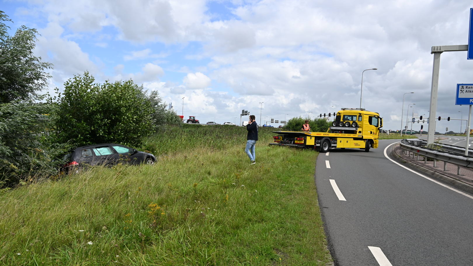 Een auto is van de weg geraakt en staat in de berm tussen hoge gras en struiken, terwijl een gele bergingsvoertuig op de weg klaarstaat om de auto te bergen.