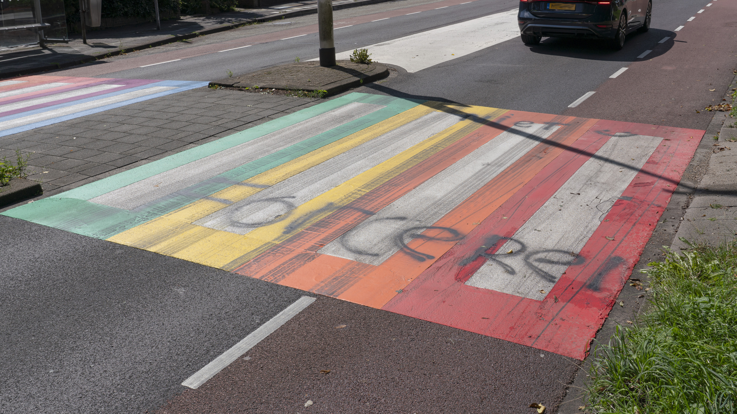 Een regenboogzebrapad met meerdere kleuren, waaronder groen, geel, rood en paars, overschilderd met graffiti, op een weg waar een auto in de verte rijdt.