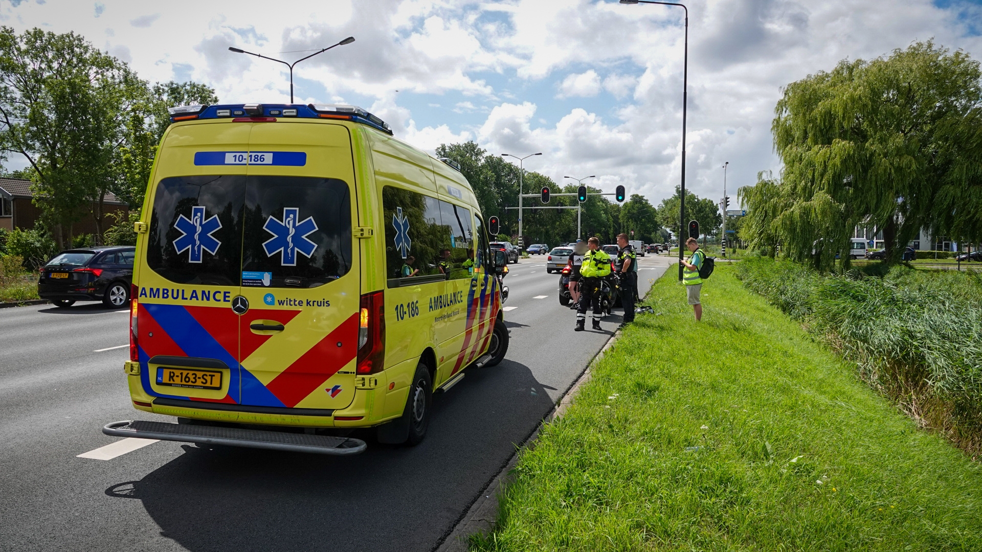 Gele ambulance geparkeerd langs een weg, met politieagenten en een persoon in een veiligheidsvest in gesprek verderop. Er zijn stoplichten en auto's op de achtergrond aanwezig, en gras en bomen aan de zijkant van de weg.