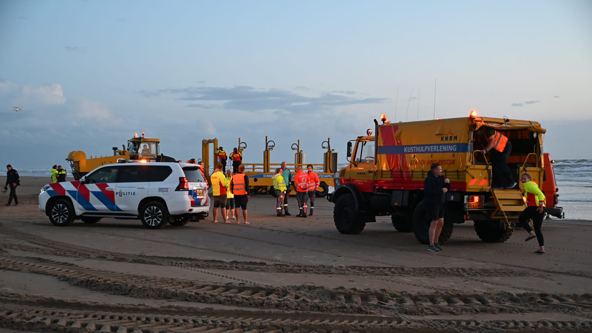 Hulpdiensten, waaronder politie en KNRM, verzamelen zich met voertuigen en personeel op een strand tijdens een avondoperatie; de zee en een vorkheftruck zijn op de achtergrond zichtbaar.