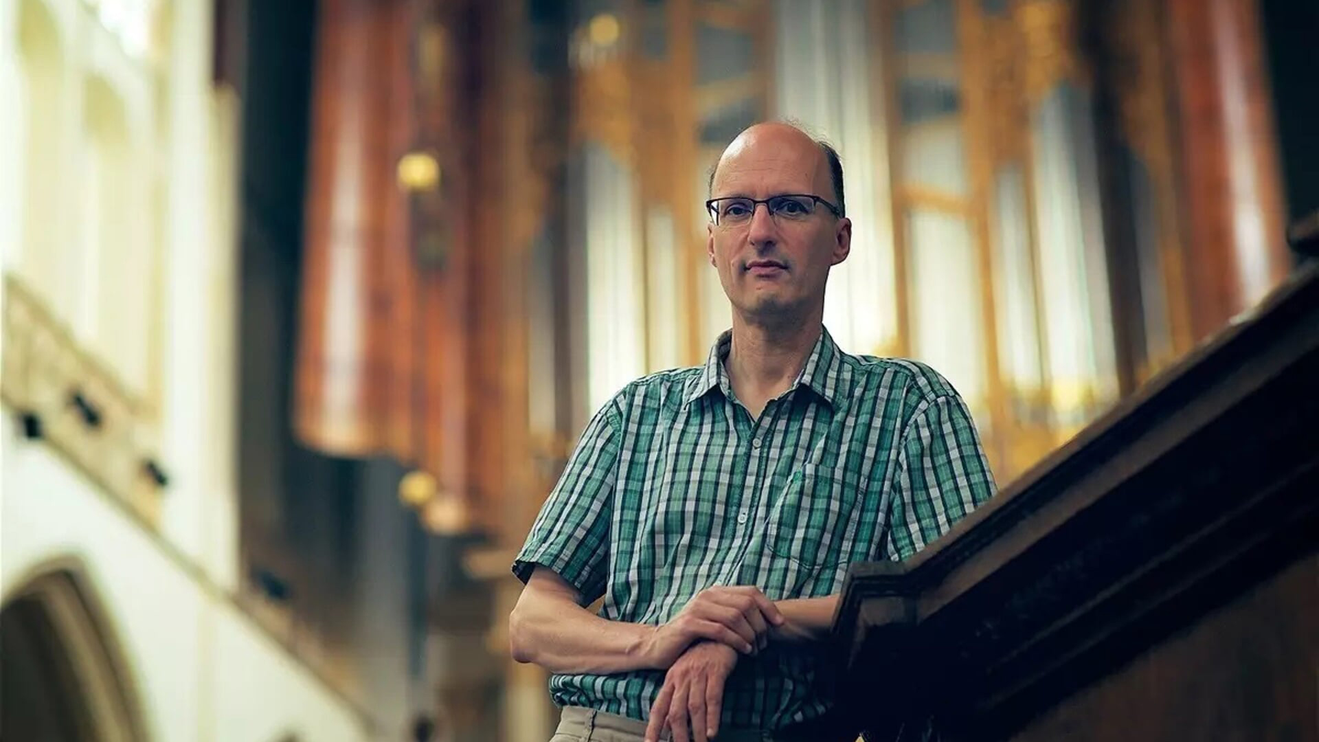 Man met bril en geruite blouse staat in een kerk, leunend op een houten balustrade met pijpenorgel op de achtergrond.