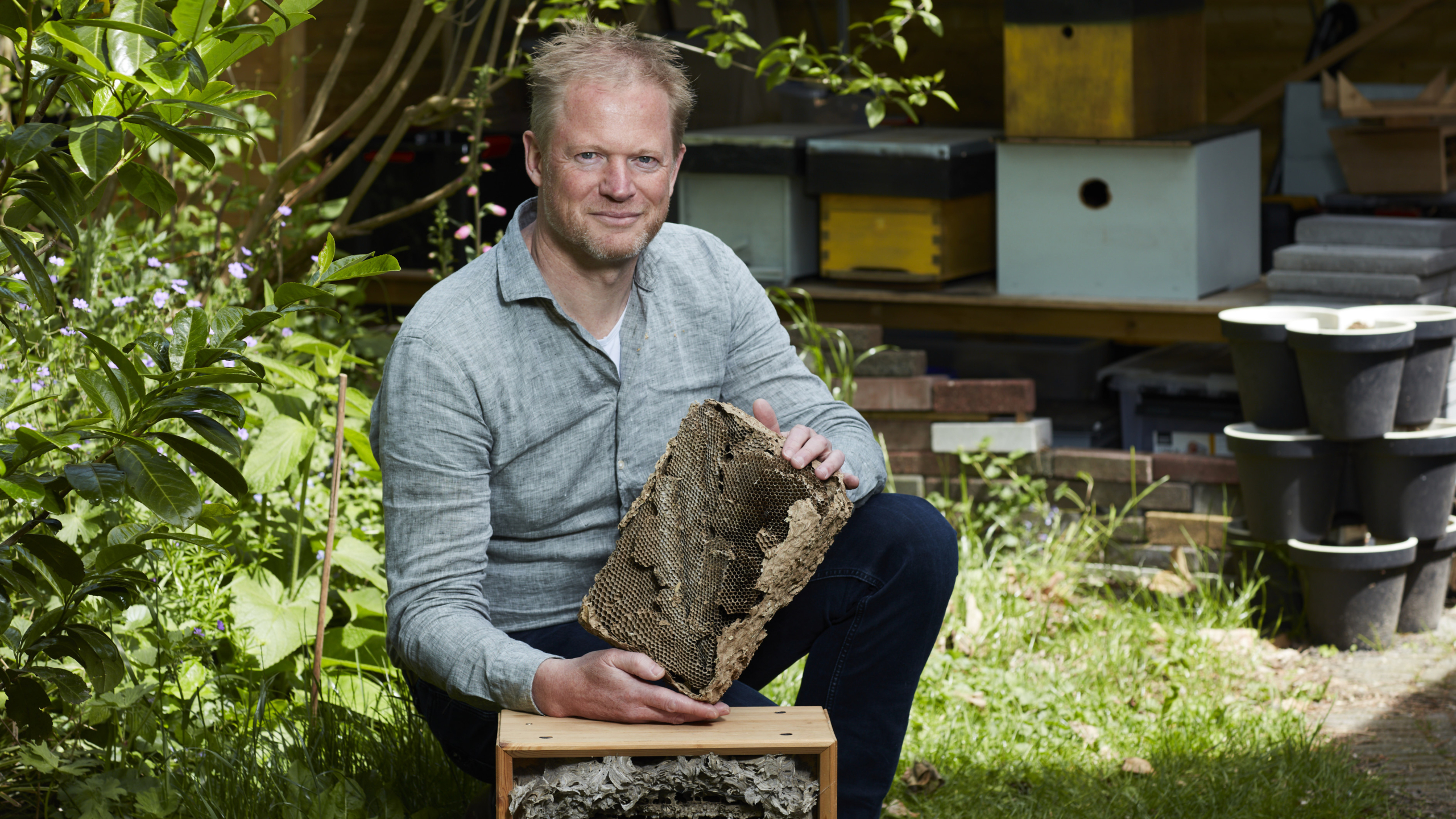Een man zit gehurkt in een tuin en houdt een stuk bijenraat vast. Op de achtergrond zijn een bijenkast en verschillende planten te zien.
