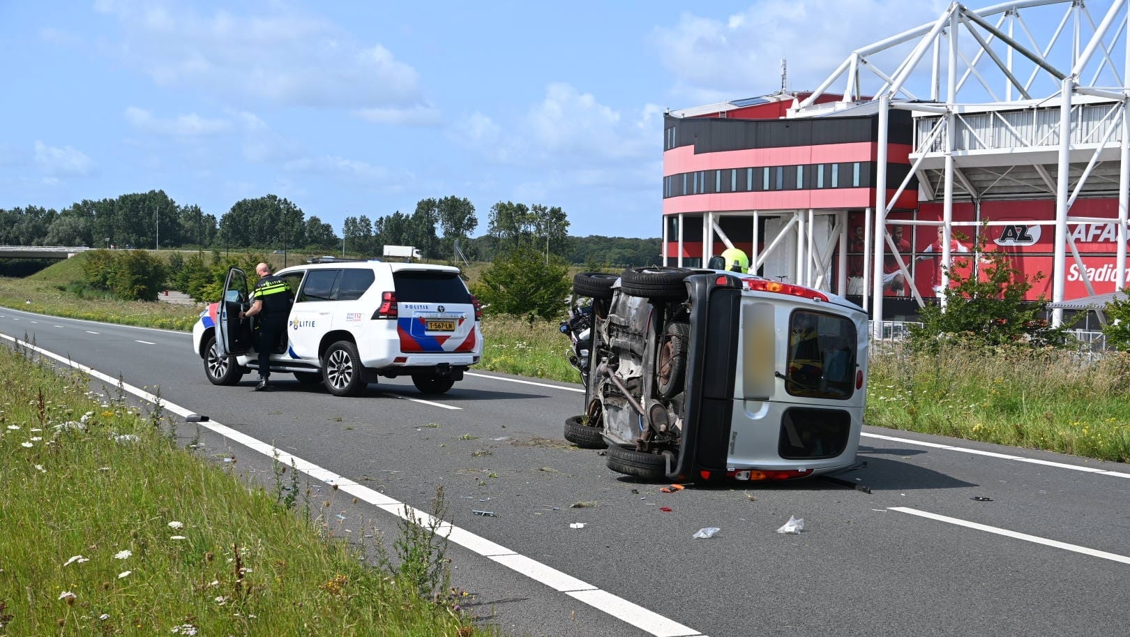 Een omgeslagen auto ligt op zijn zijkant op de weg bij het AFAS Stadion, met een politieauto en agent ter plaatse.