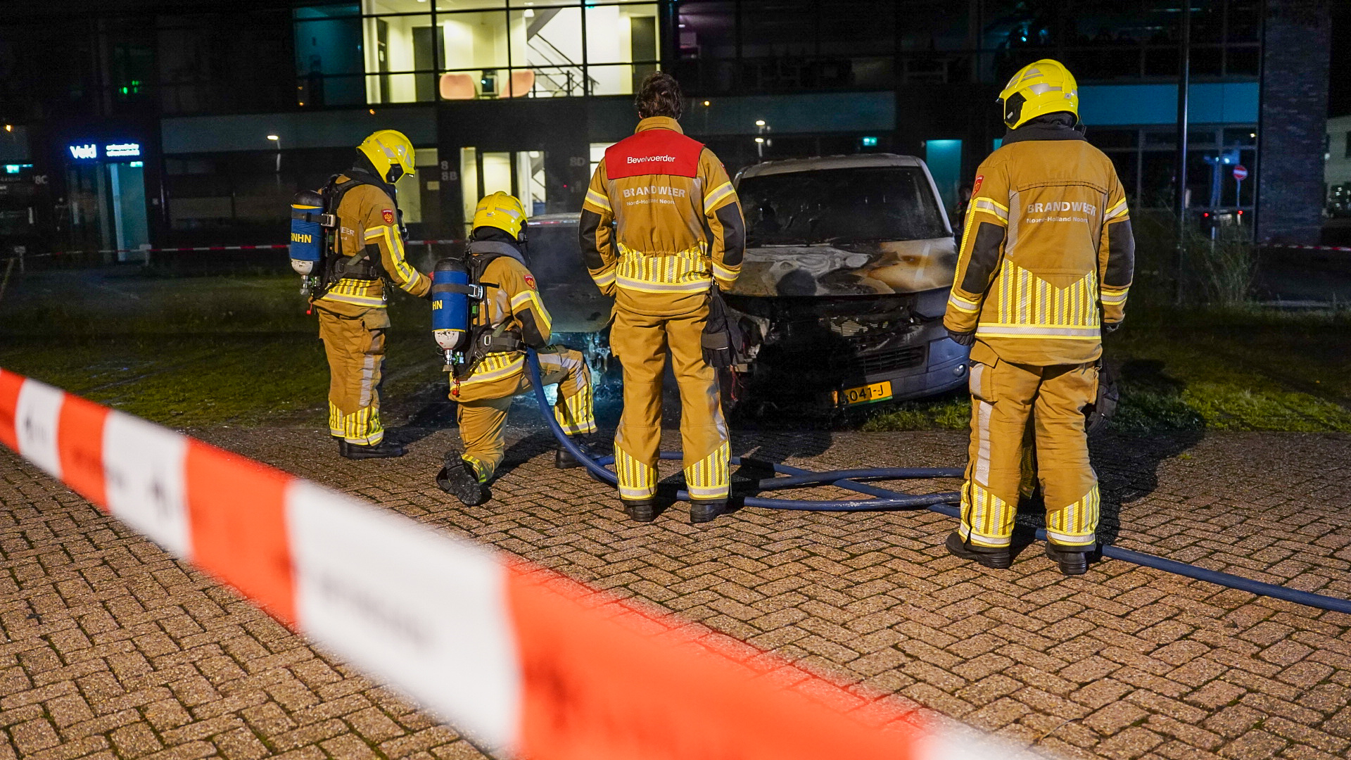 Brandweerlieden bij een gebouw met een brandende auto, afgeschermd door rood-wit lint, in de nacht.