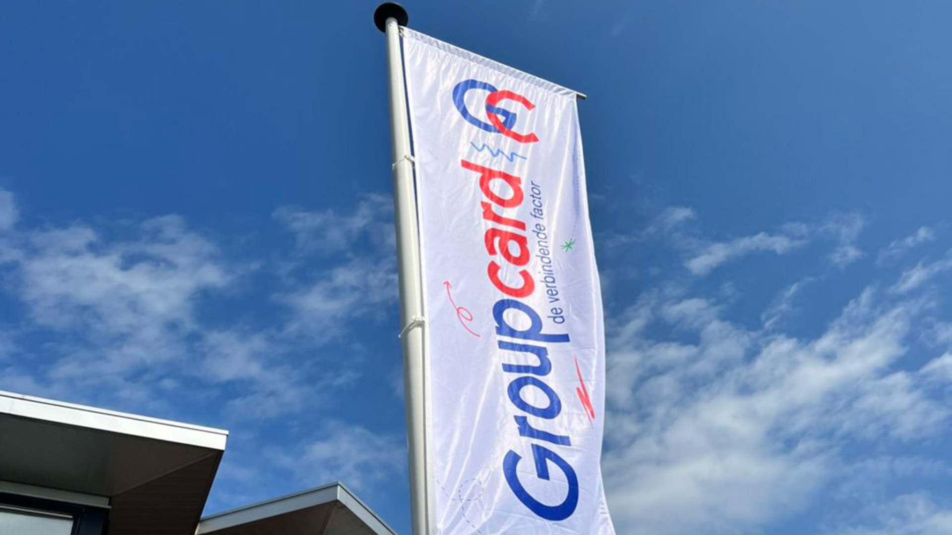 Vlag met de tekst 'Groupcard, de verbindende factor' wappert aan een vlaggenmast tegen een blauwe hemel met enkele wolken.