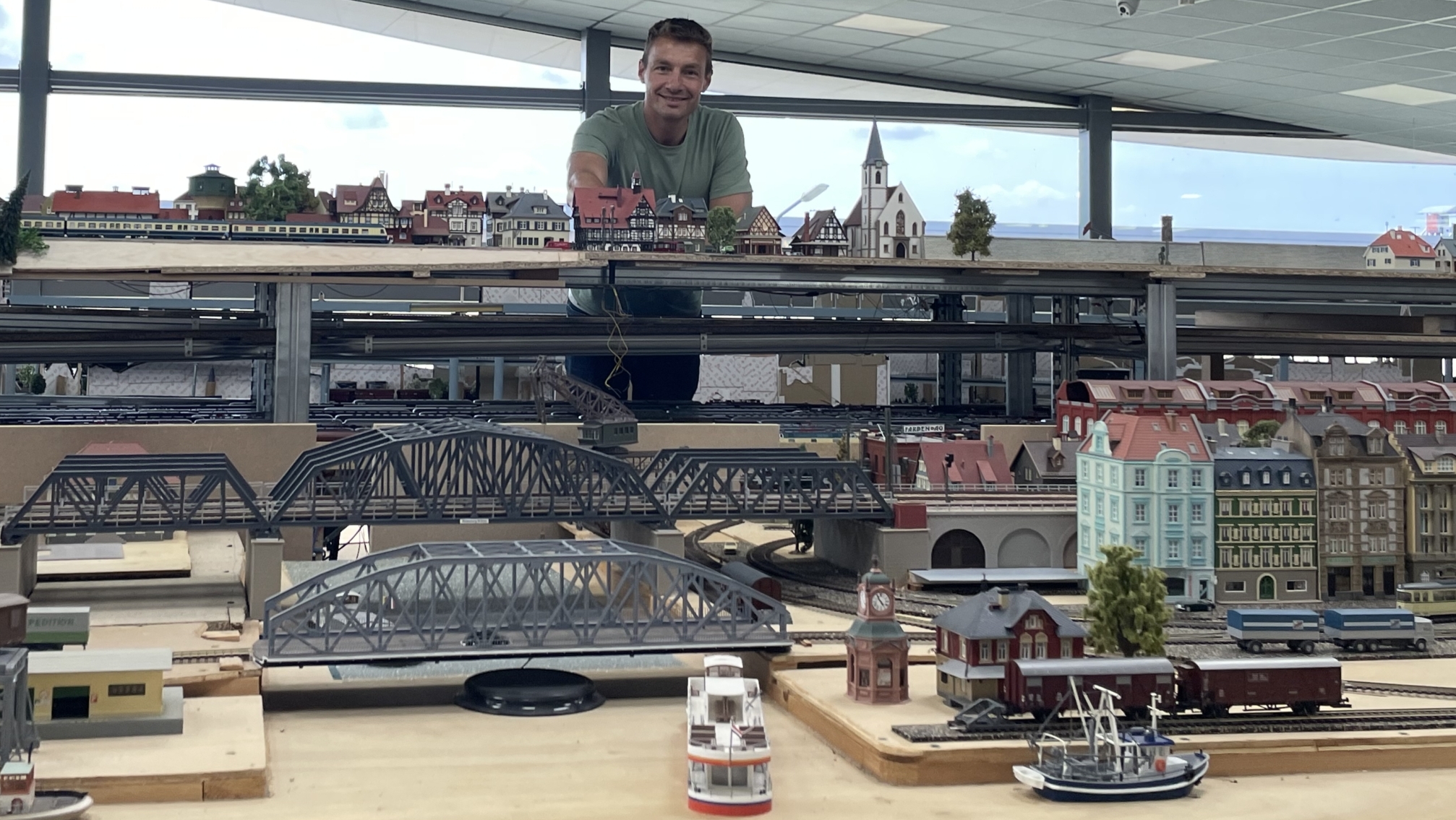 Een persoon staat achter een uitgebreide modelspoortafel met gedetailleerde miniatuurgebouwen, bruggen en treinen in een overdekte ruimte met glazen wanden.