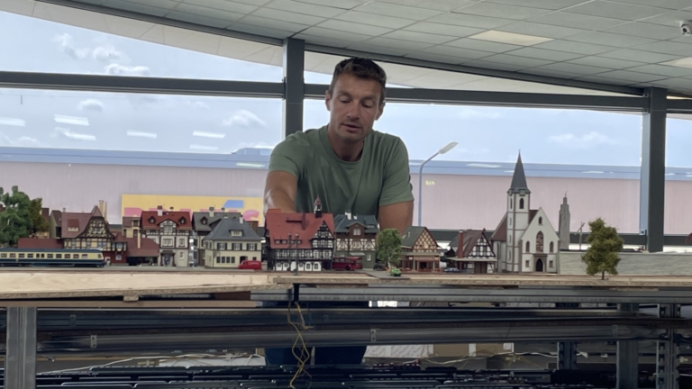 Een man in een groene T-shirt observeert een modeltrein en miniatuurstation in een grote hal met een glazen wand.
