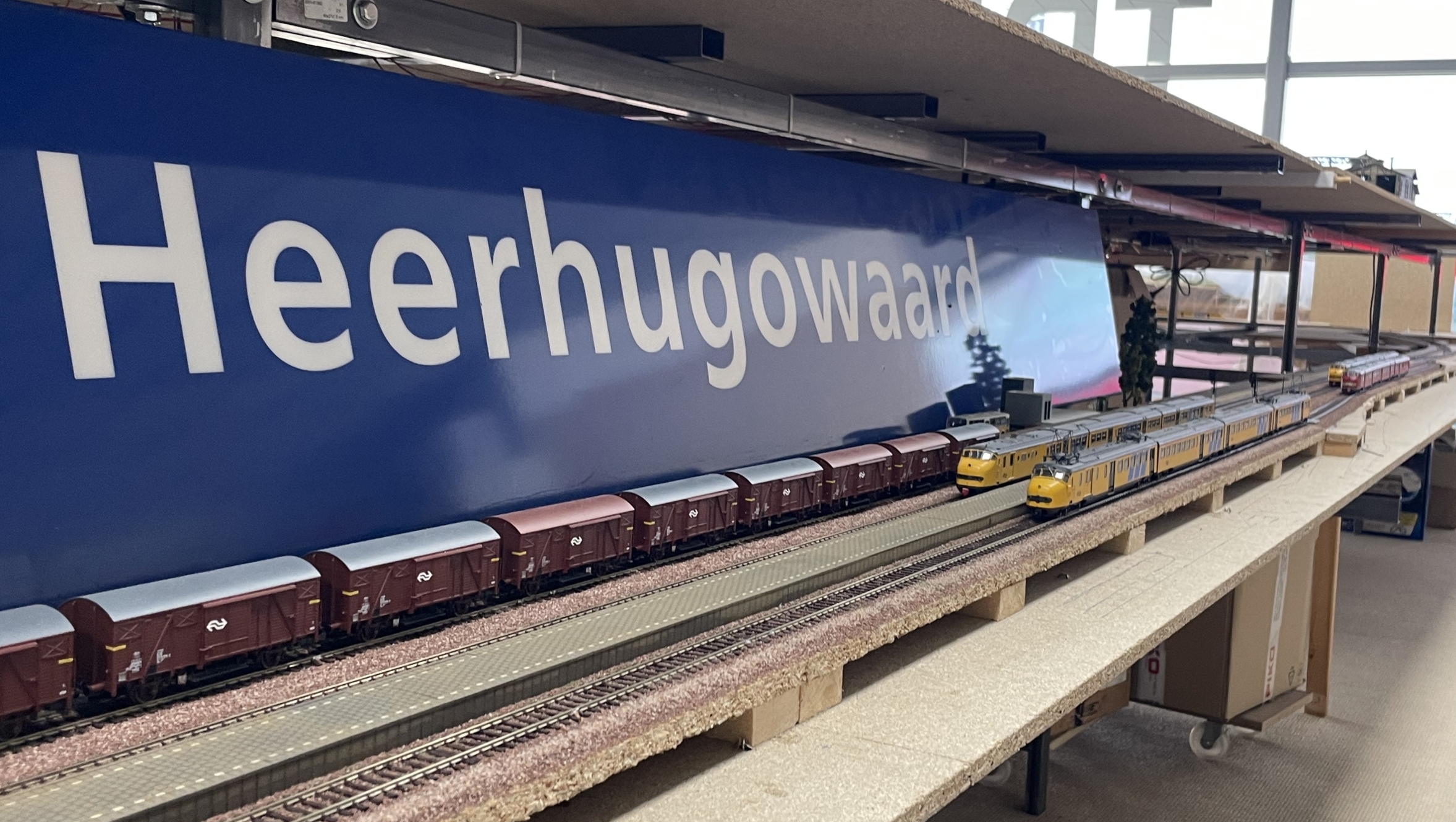 Modelspoorwegbaan met een bord 'Heerhugowaard' en miniatuurtreinen op de sporen, geplaatst in een binnenruimte met grote ramen.