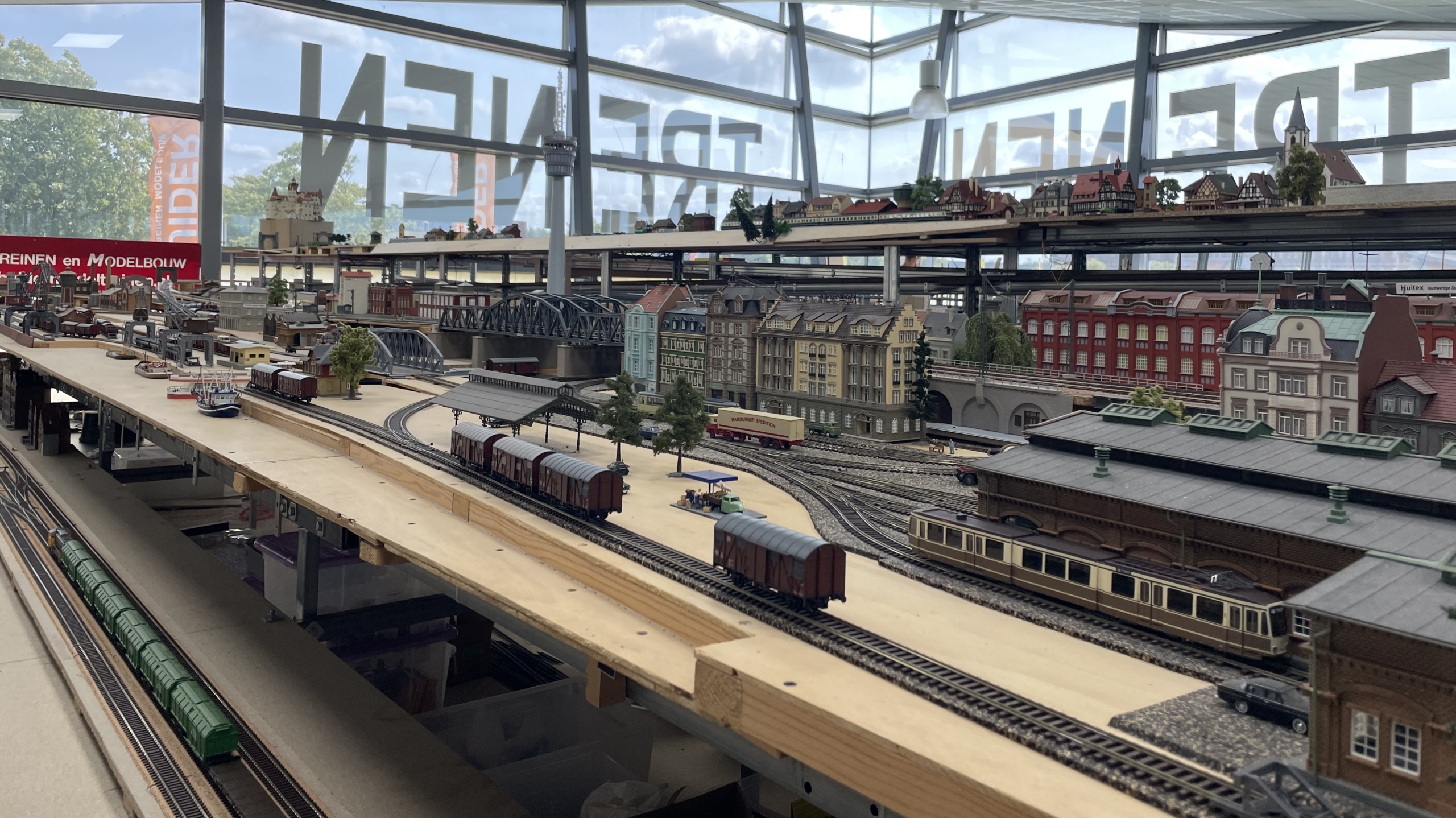 Modelspoorbaan op een tafel in een kamer met grote ramen, met miniatuurtreinen, huizen en een brug op de achtergrond.