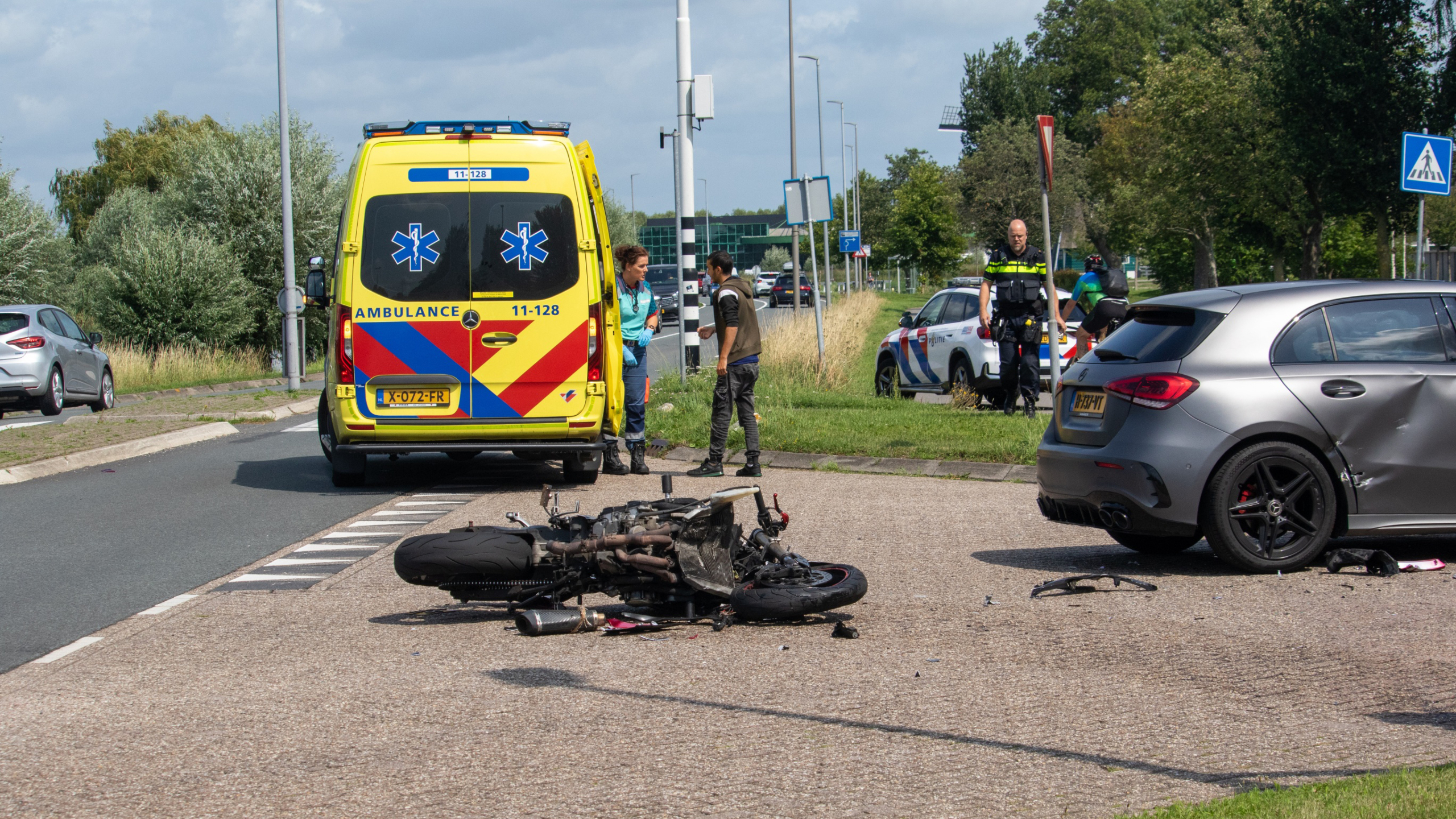 Gele ambulance en politievoertuig staan naast een beschadigde motorfiets en grijze auto op de weg, met enkele mensen die ter plaatse staan.